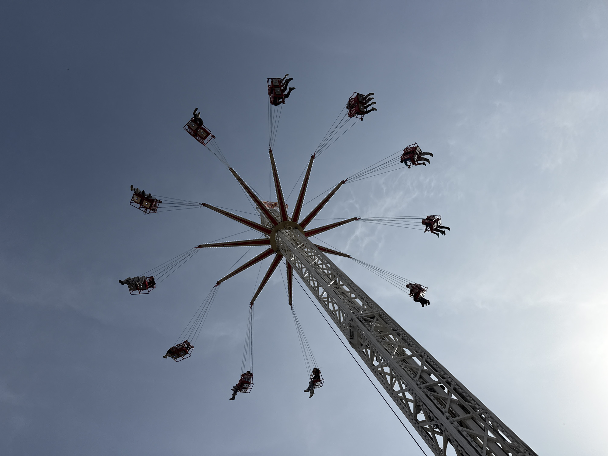 The image shows a Star Flyer amusement ride in Plovdiv, Bulgaria. The ride is tall with a central tower structure made of white lattice metal. From the top of the tower, radiating outward like the spokes of a wheel, are red metal arms with hanging swings. Each swing is occupied by one to four people. The ride is in operation, and the swings are extended outward and slightly upward from the center of the tower. The background is a light blue sky with a few faint clouds. The sun is on the right side of the frame, creating a bright area.