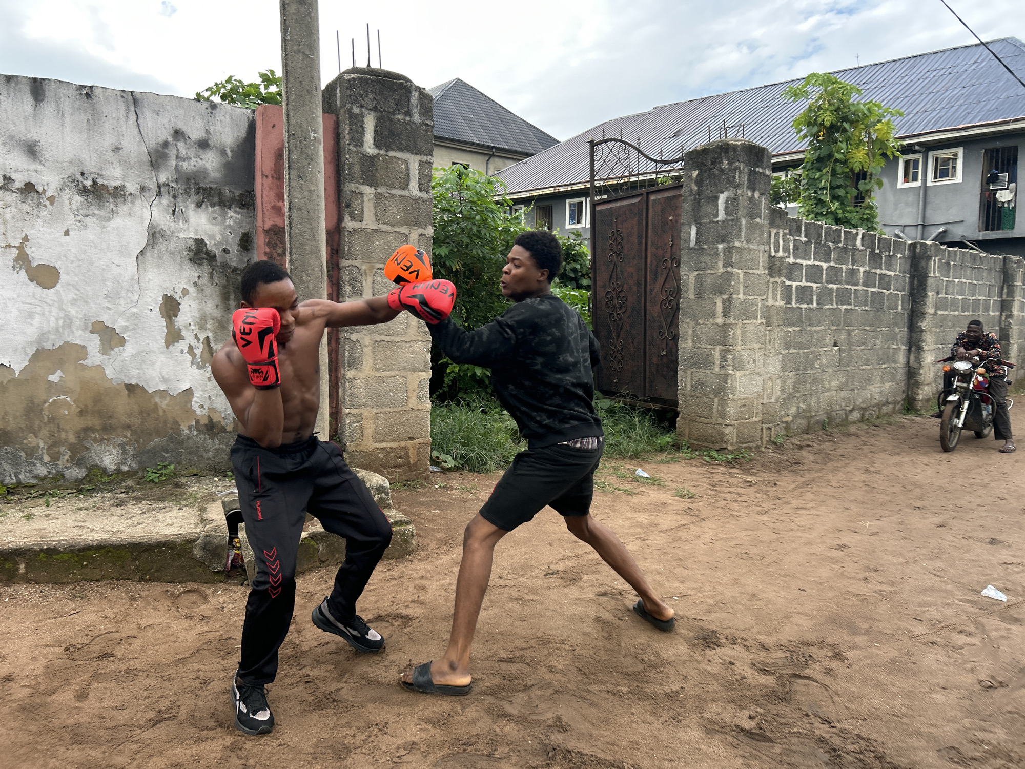 Two young men are sparring with boxing gloves outdoors on a dirt ground in what appears to be a residential area in Ihiagwa, Nigeria. The man on the left is shirtless, wearing black pants with red trim and black and white sneakers. He is throwing a punch with his right hand. The man on the right is wearing a dark long-sleeved shirt, black shorts, and black sandals. He is blocking the punch. In the background, there is a weathered concrete wall, a metal gate, and buildings with corrugated metal roofs. Further in the background, a person is riding a motorcycle. The sky is overcast.