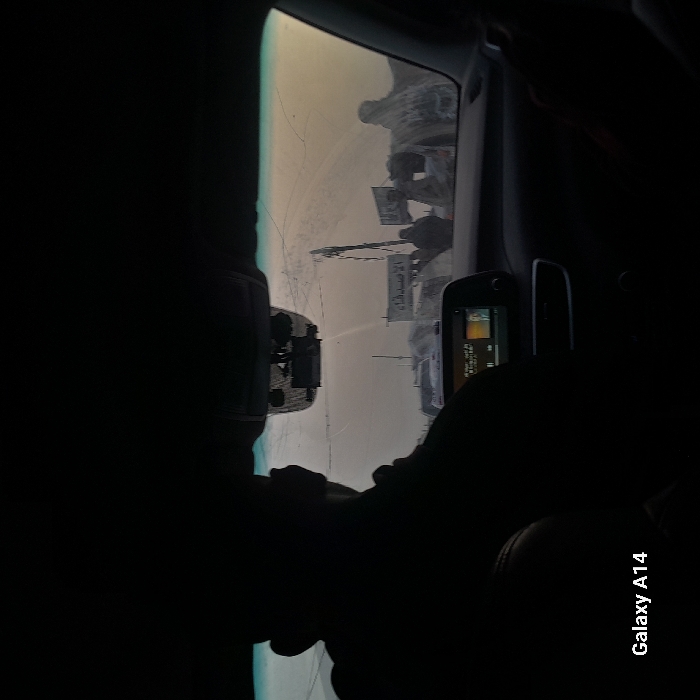 View from inside a car. In the background, visible through the front windshield, are people holding signs in what appears to be a dusty or hazy outdoor setting. There is a structure with a tower visible in the distance, possibly a minaret or other religious structure. Part of a navigation system is visible. A dark figure sits in the car in a slumped position. The "Galaxy A14" watermark appears at the bottom of the image.