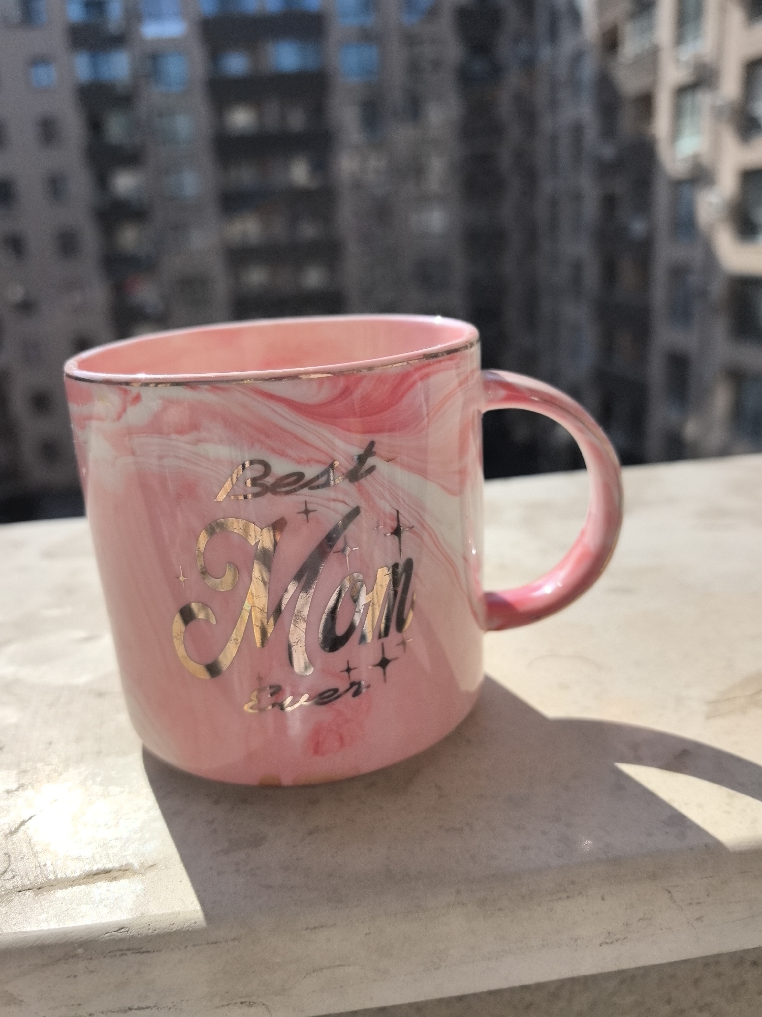 The image shows a pink mug with white marbling and gold trim. The words "Best Mom Ever" are printed on the mug in gold lettering. The mug is sitting on a light-colored surface, casting a shadow. In the background, a blurred cityscape is visible. Based on the architectural style, it may be Plovdiv, Bulgaria. No people are visible.