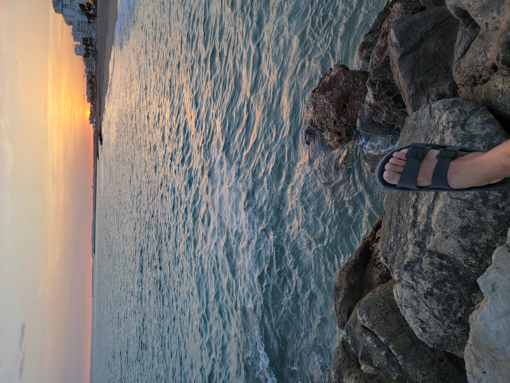 A foot wearing a dark gray sandal rests on large, rough-textured rocks that form a breakwater or embankment along the coast. The rocks vary in color from light gray to dark brown and are partially submerged by the choppy, blue-green sea. In the background, the ocean stretches to the horizon under a sky illuminated by a setting sun. The sun's warm orange and yellow hues cast a glow on the water's surface and the distant coastline, where a sandy beach is visible, lined with palm trees and white buildings that appear to be hotels or apartment complexes. The city of Quarteira, Portugal, is identifiable by its characteristic buildings and beachfront. The overall scene captures a peaceful evening at the seaside.