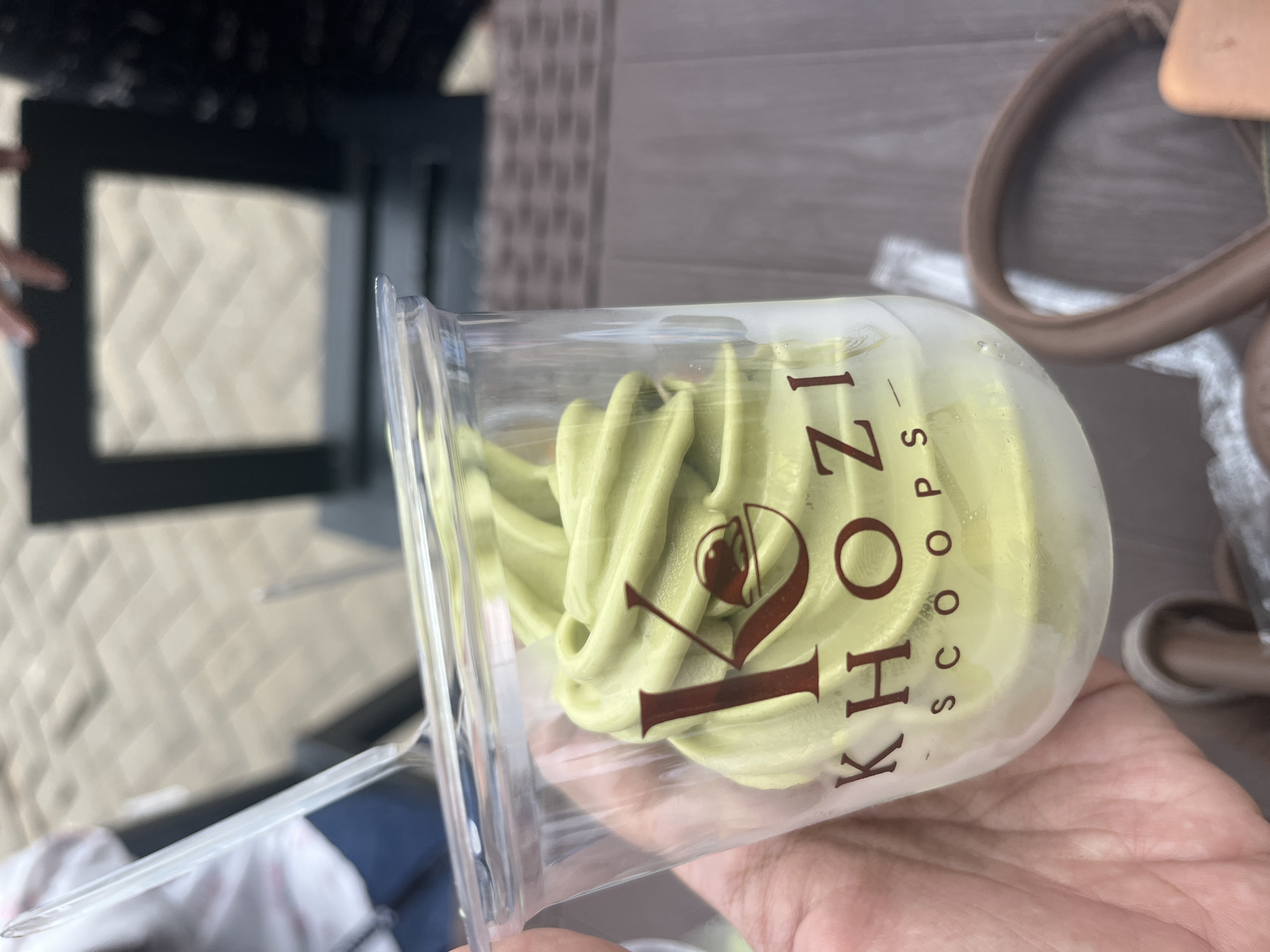 A close-up shot captures a clear plastic cup filled with vibrant green soft-serve ice cream. The cup, held by a person's hand, displays the "KHOZI SCOOPS" logo in a dark red font. The ice cream has a swirled, soft-serve texture. In the background, out of focus, a patterned brick pathway is visible, along with parts of furniture and other individuals. The setting appears to be an outdoor cafe or seating area, possibly in Meliau Hilir, Indonesia, given the context.