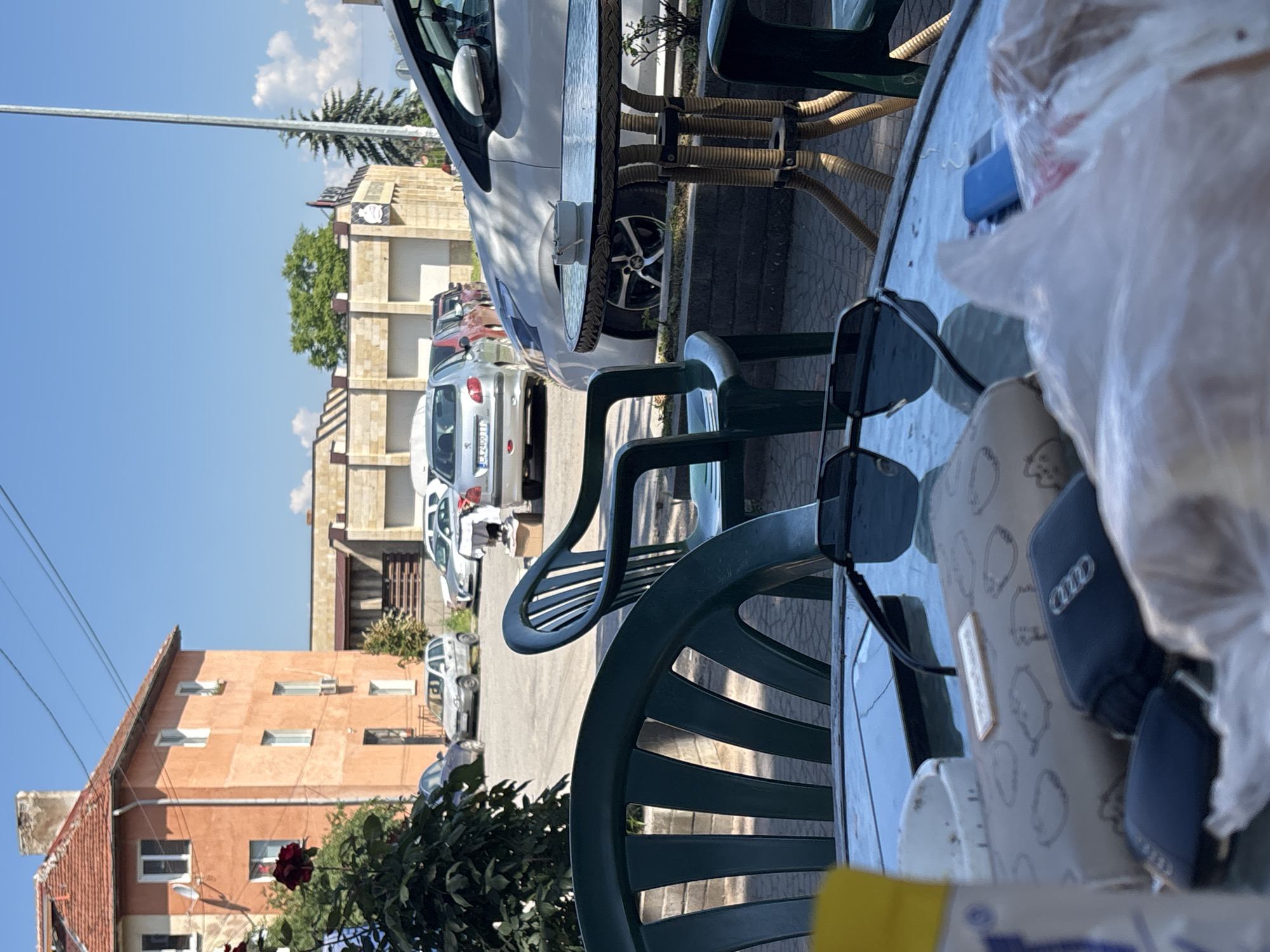 In Elin Pelin, Bulgaria, on a sunny day, an outdoor seating area is depicted. Two green plastic chairs are positioned in the foreground. In the middle ground, a glass-top table with a wicker-style base is present, with an ashtray containing cigarette butts on its surface. Several cars are parked in the background. To the left, an orange building with a red-tiled roof stands, and to the right, a light-colored building with a modern facade is visible. The sky is clear blue with some scattered clouds. On the table, a pair of sunglasses, a wallet, car keys, and a cell phone are laid out. A plastic bag is also present on the table.