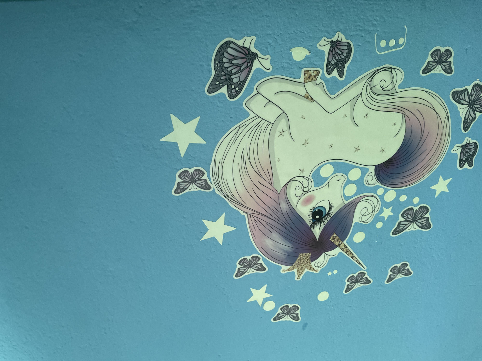 The media file displays a close-up of a blue wall decorated with stickers of a cartoon unicorn and butterflies. The unicorn, rendered in light colors with a purple mane and tail, is depicted upside down, seemingly floating amongst the butterflies and stars. Its eye is large and blue, and it has a golden horn. The butterflies are a mix of black and purple, some with intricate wing patterns. Several white stars and small white circles are also scattered across the wall, contributing to a whimsical, fantasy-themed aesthetic. The wall's textured surface is visible. The image appears to be taken indoors, possibly in a child's room or a decorated space, with artificial lighting as there are no cues for time of day or weather. There is no visible text in the image. The context of Kołobrzeg, Poland, is provided but cannot be visually inferred from the image itself.