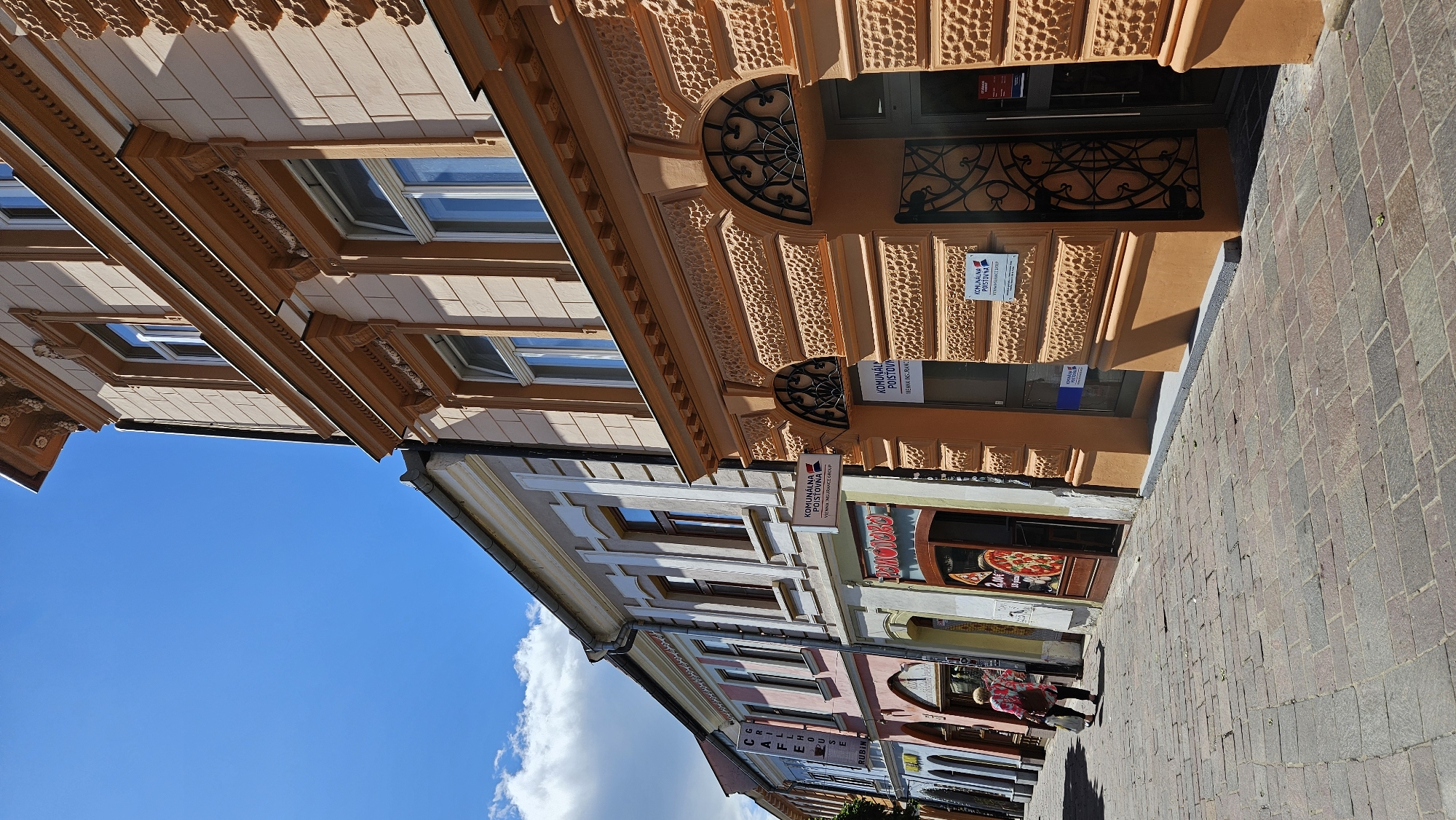 The image depicts a street scene in Prešov, Slovakia, likely on or near Hlavná Street, a prominent commercial area. The perspective is angled upwards, focusing on the facades of several buildings lining a cobblestone street. The buildings display a mix of architectural styles, with the closest one to the right exhibiting ornate orange stucco decoration, arched windows with intricate metalwork, and signage for "KOMUNÁLNA POISŤOVŇA" (Municipal Insurance) and "VIENNA INSURANCE GROUP." Further down the street, buildings in pastel colors like pink and cream are visible, featuring more traditional window designs and signage for businesses such as "CAFE GRILLHOUSE" and "RUBIN."
In the foreground, a woman walks away from the camera, carrying shopping bags. The cobblestone street is partially shaded by the buildings. The sky above is a clear blue with a few scattered white clouds. The overall atmosphere suggests a bright, sunny day in a historical urban setting.