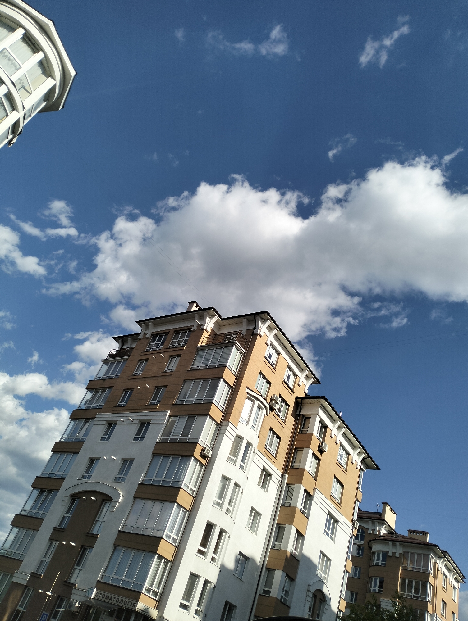 The image shows a tall, modern residential building in Krykhivtsi, Ukraine, under a blue sky with scattered white clouds. The building has a predominantly white facade with sections in a tan color, and multiple windows. A portion of another similar building is visible in the upper left corner of the frame and another on the bottom right. The building's architectural style suggests it is a relatively new construction. At the building's base, a sign with Cyrillic text, translated "Stomatology", indicates a dental clinic located within the building. The low angle of the shot emphasizes the building's height. There are no people visible in the image.