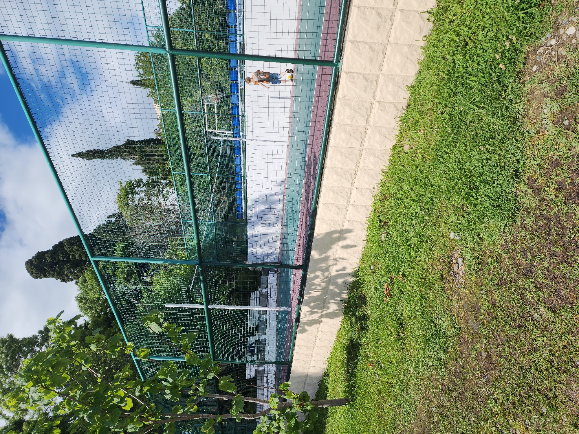 The image shows a person standing on a tennis court in Sochi, Russia. The court is enclosed by a green fence and is located next to a grassy area and a sidewalk. The weather appears to be sunny with some clouds in the sky.