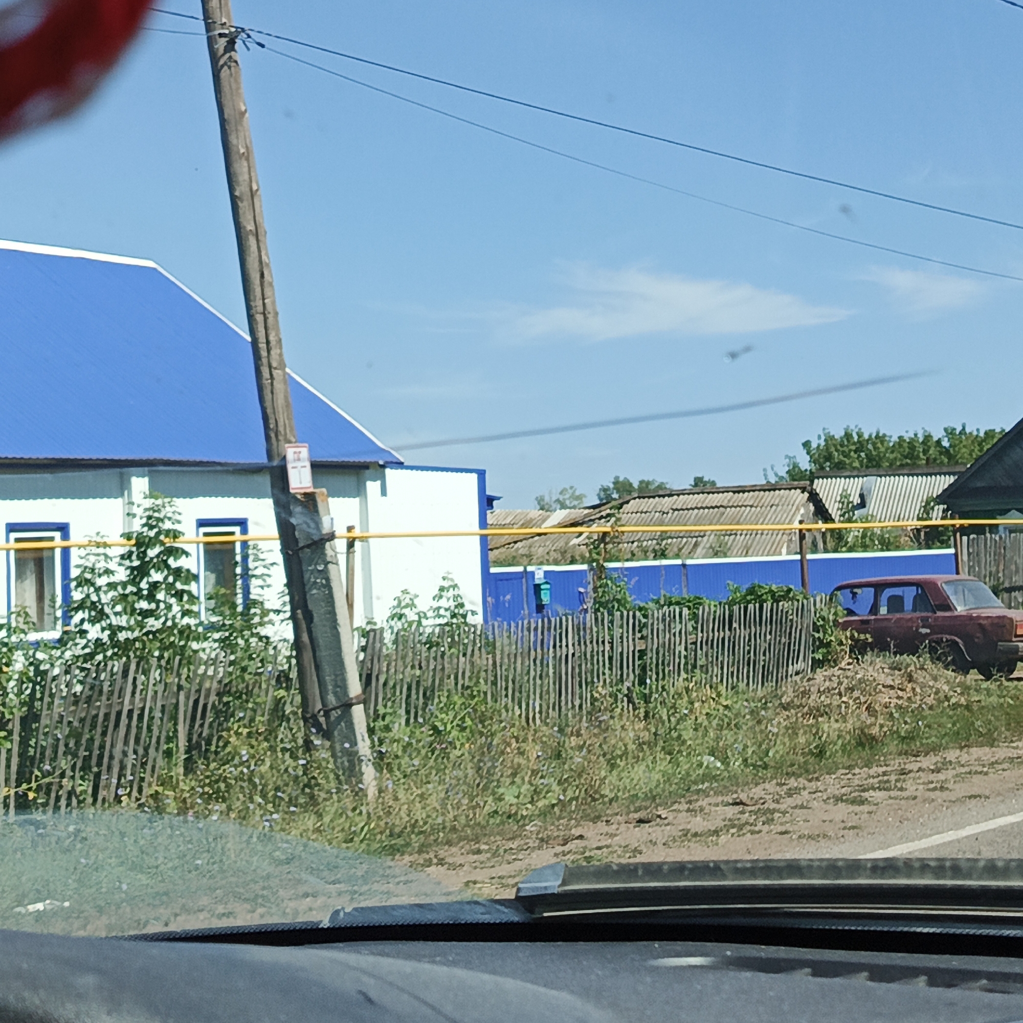 A rustic scene unfolds under a clear blue sky in the village of Raznomoyka, Russia. A weathered wooden utility pole stands prominently on the left, adorned with a small sign displaying an "H" symbol. Behind it, a white building with a bright blue roof features two windows with blue trim and a yellow pipe running horizontally across its facade. A rustic wooden fence separates the property from a grassy area, beyond which lies a blue fence and further structures. A faded red car is parked to the right, its windows dark. The ground is a mixture of dirt and dry grass, with some green foliage scattered throughout. The foreground shows the interior of a vehicle, with a dark dashboard and windshield.