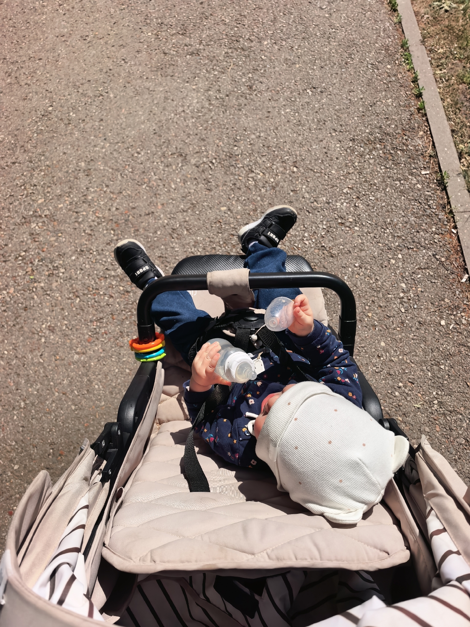 A top-down view shows a baby in a stroller, holding a baby bottle. The baby is wearing a patterned navy blue long-sleeved shirt, jeans, and black shoes with the word "SPORT" printed on them, and a white cap. The stroller is beige and the baby is strapped in with a black harness. The stroller is on a gray paved road next to a patch of green grass near a curb. The scene takes place in Sterlitamak, Russia.