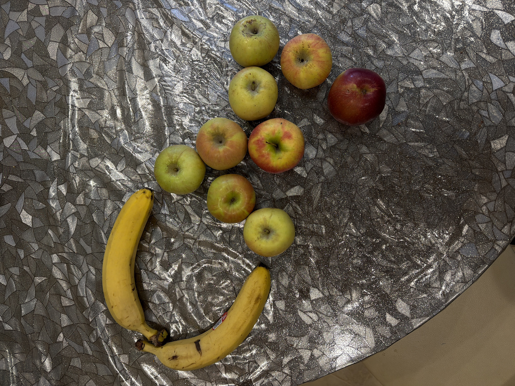 On a table with a silver patterned surface are arranged a cluster of ten apples and two bananas. The apples display a range of colors, from green to yellow with red hues. The bananas are yellow and appear to be slightly bruised. The location is unspecified within the city of Kamyshin, Russia, as no location details are present in the image.