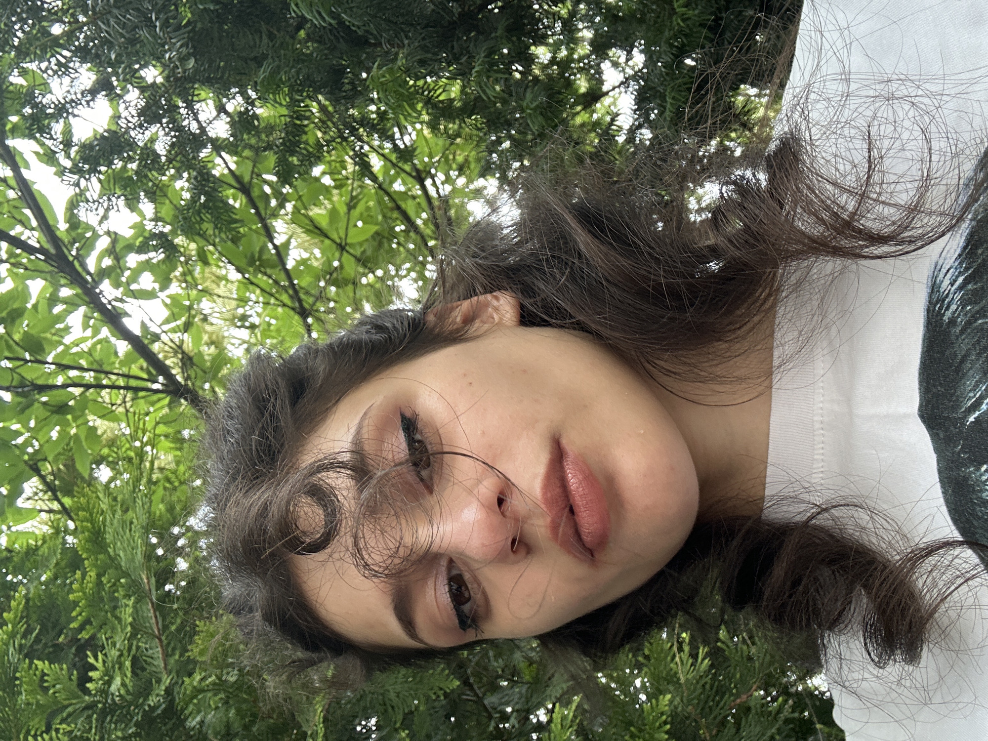 A low-angle, close-up selfie shows a young woman with dark, curly hair framing her face. She is outdoors, with lush green trees and foliage visible in the background, suggesting a park or garden setting. The woman is wearing a white t-shirt with a dark graphic on the front. Her gaze is directed towards the camera, with subtle eye makeup and a soft lip color. The lighting suggests it is daytime. The location is not specified.