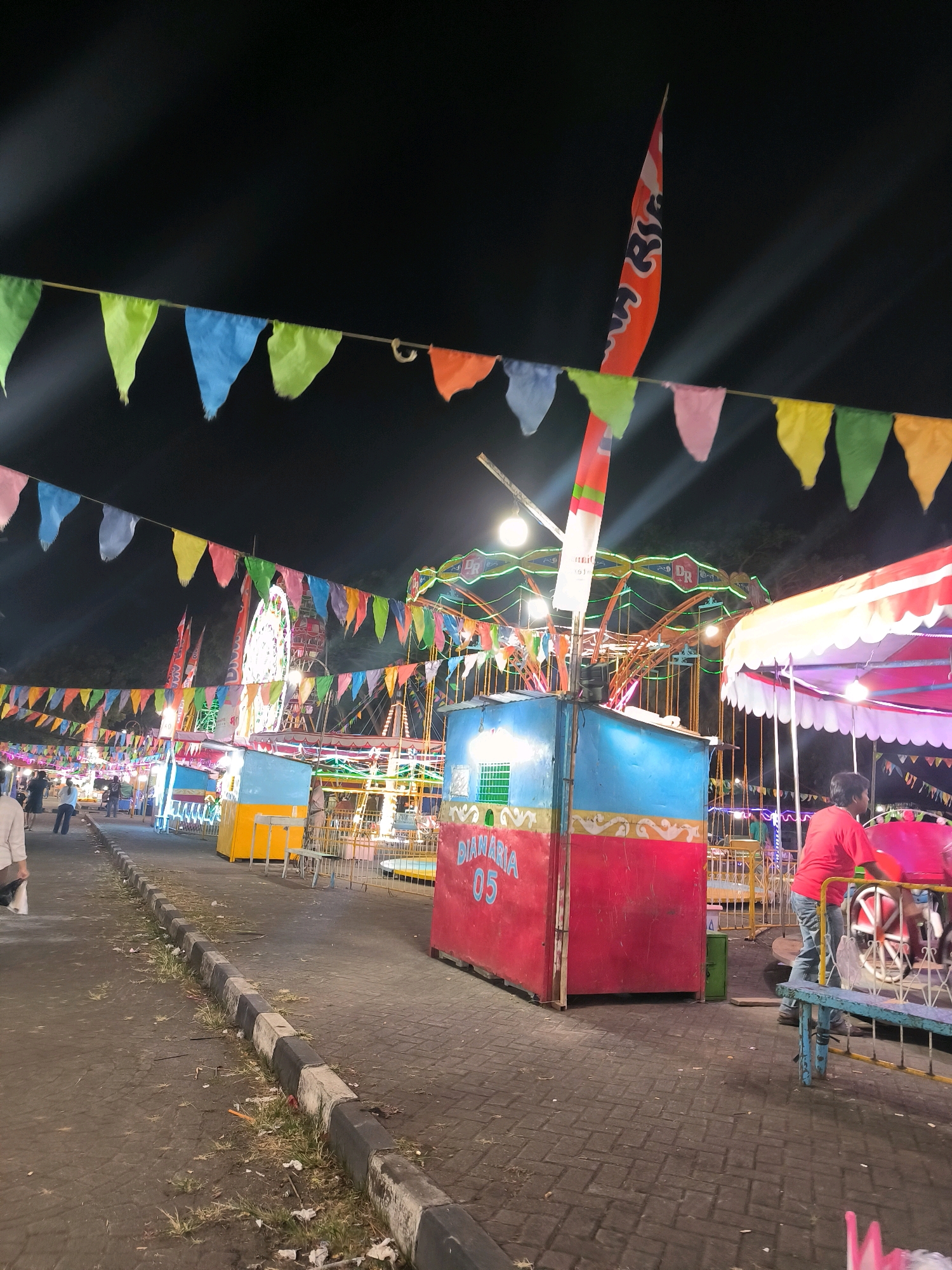 This is a nighttime photograph of a small amusement area in Ambarawa, Indonesia. Colorful pennant flags are strung across the scene, with a large orange and white flag featuring Japanese text prominently displayed on the right. In the foreground, a person in a red shirt and blue jeans is seen from the side, operating or standing next to a carnival ride. To the left, a red and blue booth with the text "DIAMARIA 05" is visible. Further in the background, more carnival rides, including a carousel-like attraction and what appears to be a Ferris wheel, are illuminated and operational. The ground is paved with bricks, and a curb with some debris runs along the left side of the frame. The overall atmosphere suggests a lively, community event.