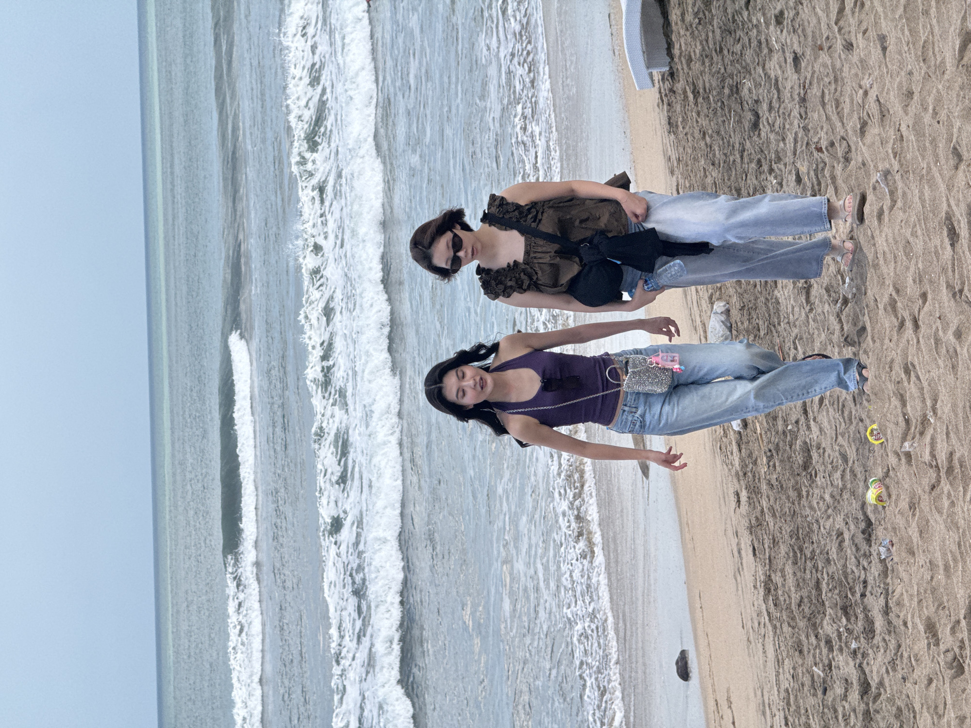 Two women walk along the beach in Kuta, Indonesia. The woman on the left has long dark hair and wears a purple tank top and light-wash jeans. She carries a small silver beaded bag. The woman on the right has short dark hair, wears a brown ruffled top, and light-wash jeans. She has sunglasses on and carries a black bag over her shoulder and a water bottle in her hand. They are walking on the sandy beach with the ocean and waves in the background. There are a few pieces of trash on the sand near the women.