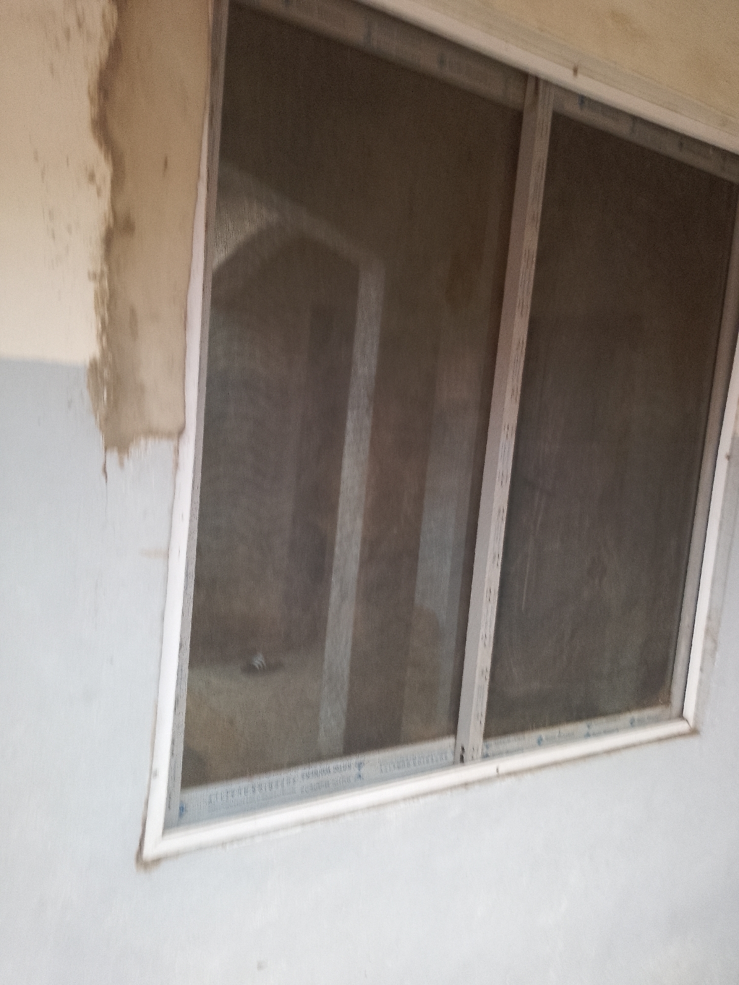 A close-up shot captures a section of an exterior wall featuring a two-panel window with a screen. The wall to the left of the window is roughly textured and appears to be unpainted or partially plastered, with areas of exposed brick or concrete. The window frame is white, and the two panels are separated by a vertical white support. The window screens are visible, showing a dark mesh. Beyond the screens, the interior appears dimly lit and indistinct. The lower part of the wall is painted a lighter color, possibly white or light grey, and shows some rough texture. The image provides no information about people or any specific location within Jauro Yinu, Nigeria.