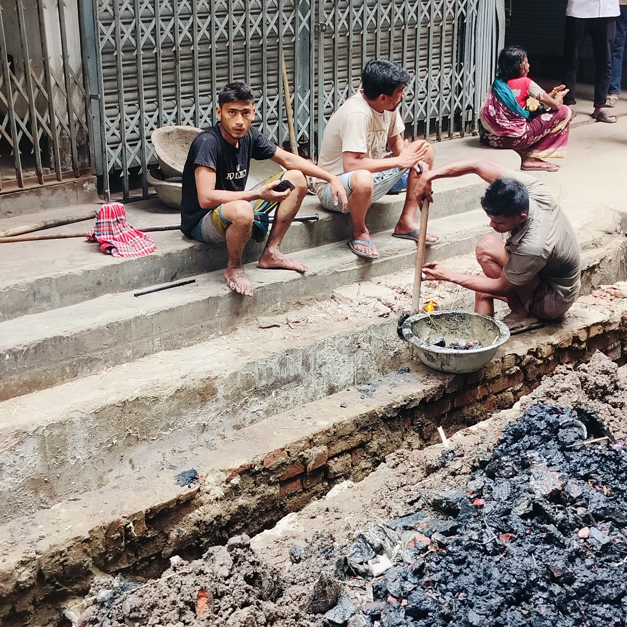 In a street scene, likely in Dhaka, Bangladesh, three men are squatting and sitting on concrete steps beside a trench being dug in the ground, filled with dark soil and brick fragments. Two of the men are sitting on the steps, while the third squats at the edge of the trench, using a pole to stir a bowl of dark material. A woman in a colorful sari and another person in white pants stand in the background. Metal gates and doors are visible behind the men. A checkered cloth lies on the steps. The man in the front is holding a mobile phone. The men are likely engaged in construction or repair work.