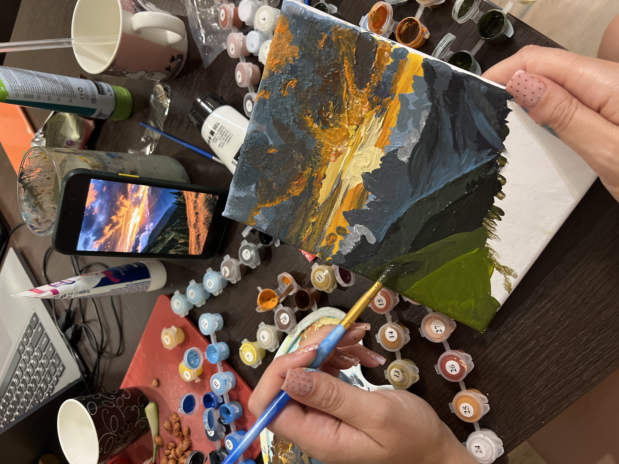 A person is painting a landscape on a canvas, using the "paint by numbers" technique. The partially completed painting, depicting a sunset or sunrise over a mountain range, is held in the person's right hand, while their left hand holds a paintbrush and dips it into a numbered paint container. The colors on the canvas include shades of blue, grey, yellow, and green.
Scattered around the canvas and paint containers are various objects: a smartphone displaying a similar sunset landscape, paint tubes, cups, a container of peanuts, and a laptop. The paint containers are labeled with numbers corresponding to areas on the canvas.
The painting and the surrounding objects rest on a dark wooden table.