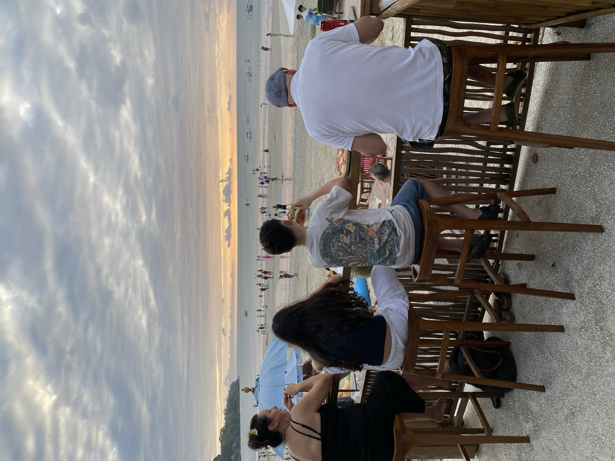 In Kuta Selatan, Indonesia, a group of four individuals is seated on bar stools at a beachfront establishment, facing away from the camera towards a scenic view of the ocean and beach at dusk. The sky is filled with overcast clouds, with a soft glow of sunset visible near the horizon. The beach is populated with numerous people, and several boats are visible on the water.
On the left, a woman wearing a black sleeveless dress is seated, with a blue umbrella visible behind her. Next to her, a person with long dark hair, wearing a dark top and white pants, is also seated. In the center, a young person in a white graphic t-shirt and denim shorts is holding a hamburger. To the right, a man in a white polo shirt and patterned shorts, wearing a grey cap, is seated. In the foreground, a pizza is visible on the table in front of the young person, and the bar area features wooden railings and tables. The ground is a patterned concrete surface.