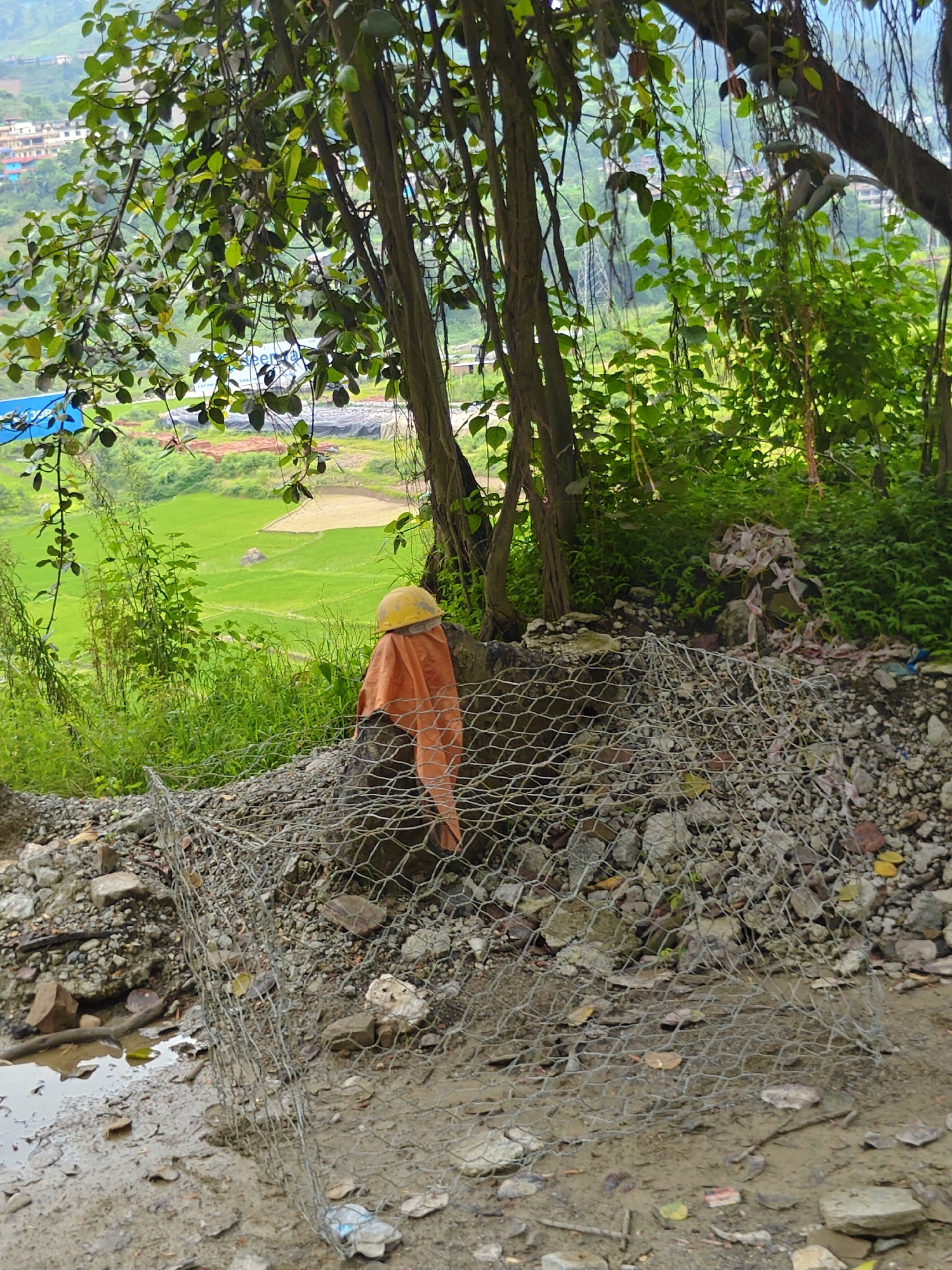 In Thakre, Nepal, a scene unfolds near a lush green landscape. In the foreground, a pile of rubble is partially covered by a wire mesh structure, suggesting ongoing construction or erosion control. A yellow hard hat rests atop an orange cloth draped over a rock, resembling a scarecrow. The background reveals terraced fields, typical of agricultural areas in Nepal, with buildings visible in the distance. Trees with dense foliage frame the scene, contributing to a natural and somewhat untamed environment. The ground is muddy and wet, indicating recent rainfall. The overall impression is one of a developing area where nature and human activity coexist.