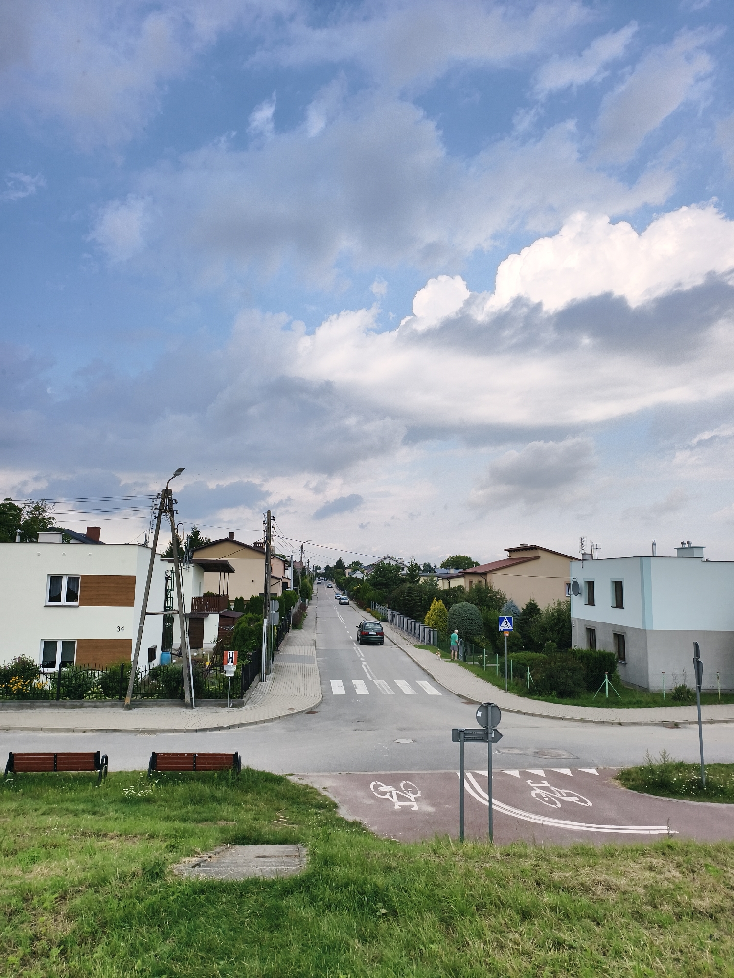 The image depicts a street scene in Tczew, Poland, featuring a residential area with houses, a road, and a grassy embankment in the foreground. The sky is partly cloudy with a mix of blue sky and white and gray clouds. Two wooden benches are situated on the grassy area. A paved road with a crosswalk runs through the scene, with a few cars visible. Several houses, varying in color and architectural style, line the street, interspersed with trees and greenery. Utility poles with electrical wires are also present. A pedestrian crossing is marked on the road. There are signs and a designated bike lane marked on the asphalt. A person in a light-colored shirt and shorts is walking a dog on the sidewalk. The overall environment suggests a suburban or residential neighborhood in Tczew.