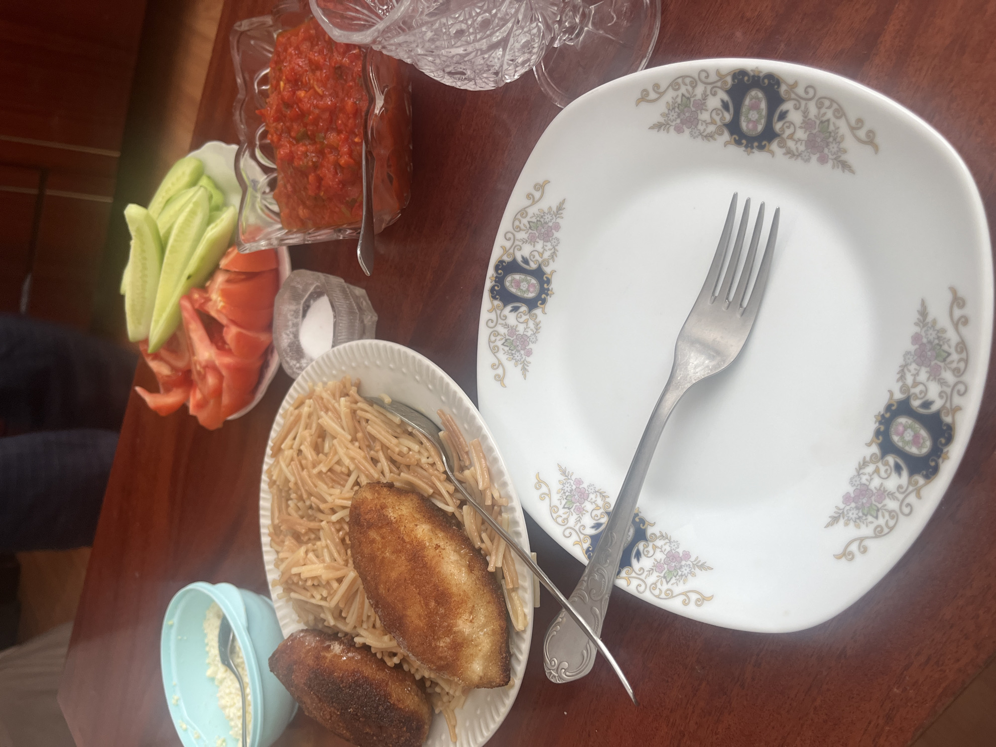 A dark wood table is set with various dishes in Yeghegnadzor, Armenia. On the left, a small white bowl contains what appears to be cooked noodles and two fried patties. A silver spoon and fork rest on the noodles and on a white plate decorated with a blue and floral pattern. The plate is empty. Adjacent to the noodles, a light blue bowl contains a white, grainy substance. Near the top left, a glass bowl holds sliced cucumbers and tomatoes. Next to this, a clear glass bowl contains a red, chunky sauce. A decorative glass object is placed next to this bowl.
