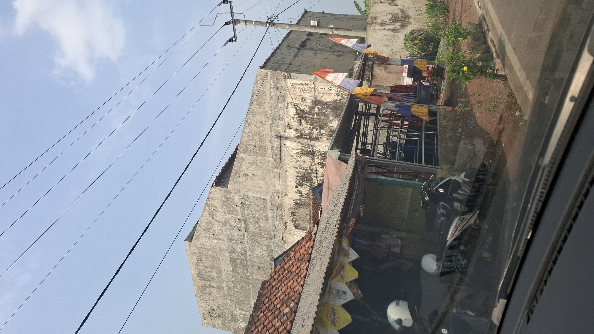 In Cibinong Hilir, Indonesia, an outdoor scene depicts a roadside view dominated by electrical wires stretching across a clear blue sky. In the mid-ground, a large, weathered concrete building stands prominently, its facade marked by age and discoloration. In front of this structure, a smaller, tiled-roof building with a corrugated metal overhang appears to be a roadside stall or shop. Flags, some with red and white patterns, are attached to poles near the stall. A motorcycle, a white helmet, and another helmet are visible in the foreground, suggesting human presence or recent activity. The ground appears to be a mix of dirt and pavement, with some vegetation and a concrete barrier. The scene captures a slice of daily life in a semi-urban Indonesian setting.