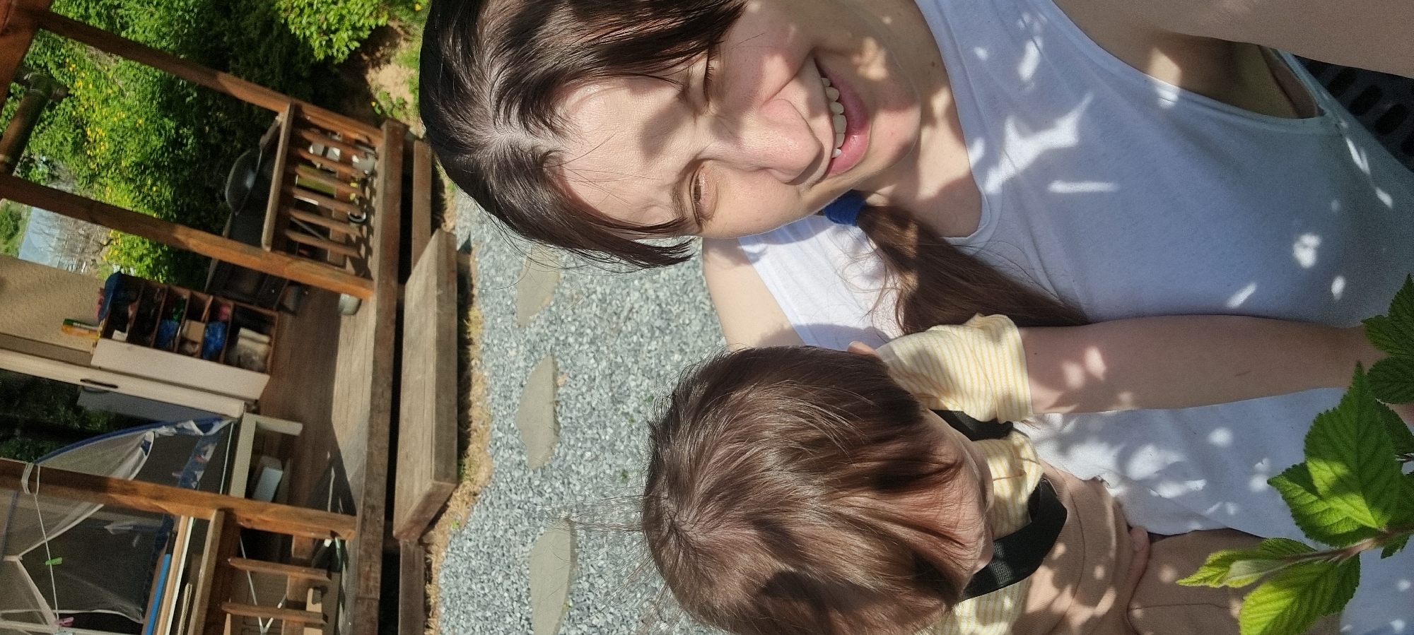 A person, likely female based on facial features and hair, is holding a baby outdoors. The person is wearing a white tank top and has brown hair tied back with a blue hair tie. The baby is wearing a light yellow and white striped shirt and a black harness. They are on a gravel path with stone steps leading to a wooden deck on the left of the frame. There's a wooden structure on the deck with various items stored within it. Green foliage is visible on the right. The location is likely Dunnae-myeon, South Korea.