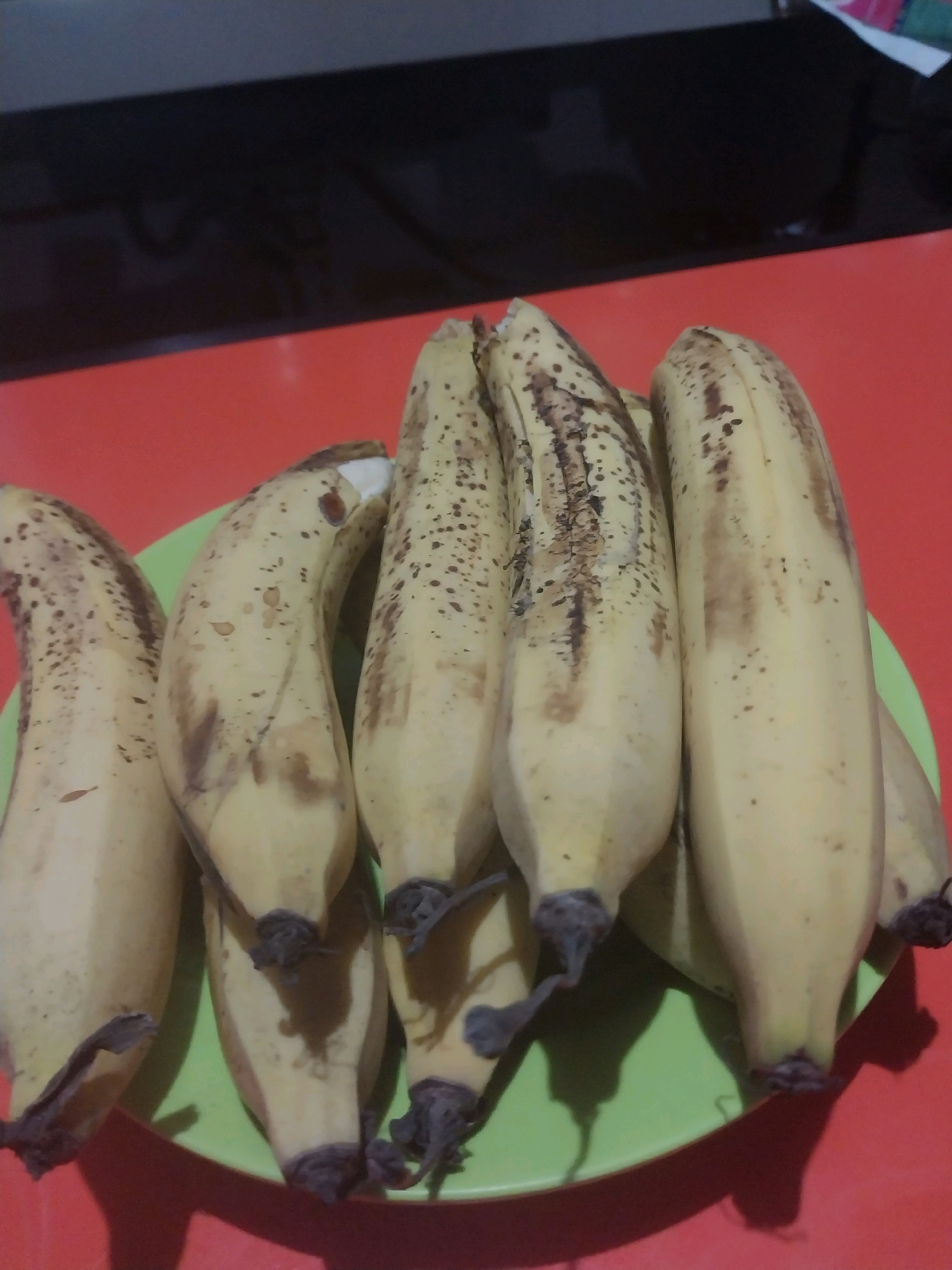 In a Blitar, Indonesia home, a bunch of six ripe bananas with brown spots sits on a bright green plate on a red surface. The bananas are oriented with their stems pointing downwards and are arranged in two rows, with three in the front and three behind them, slightly overlapping. The lighting in the scene is dim, casting shadows from the bananas onto the plate and the red surface. The background is dark and out of focus, suggesting an indoor setting.