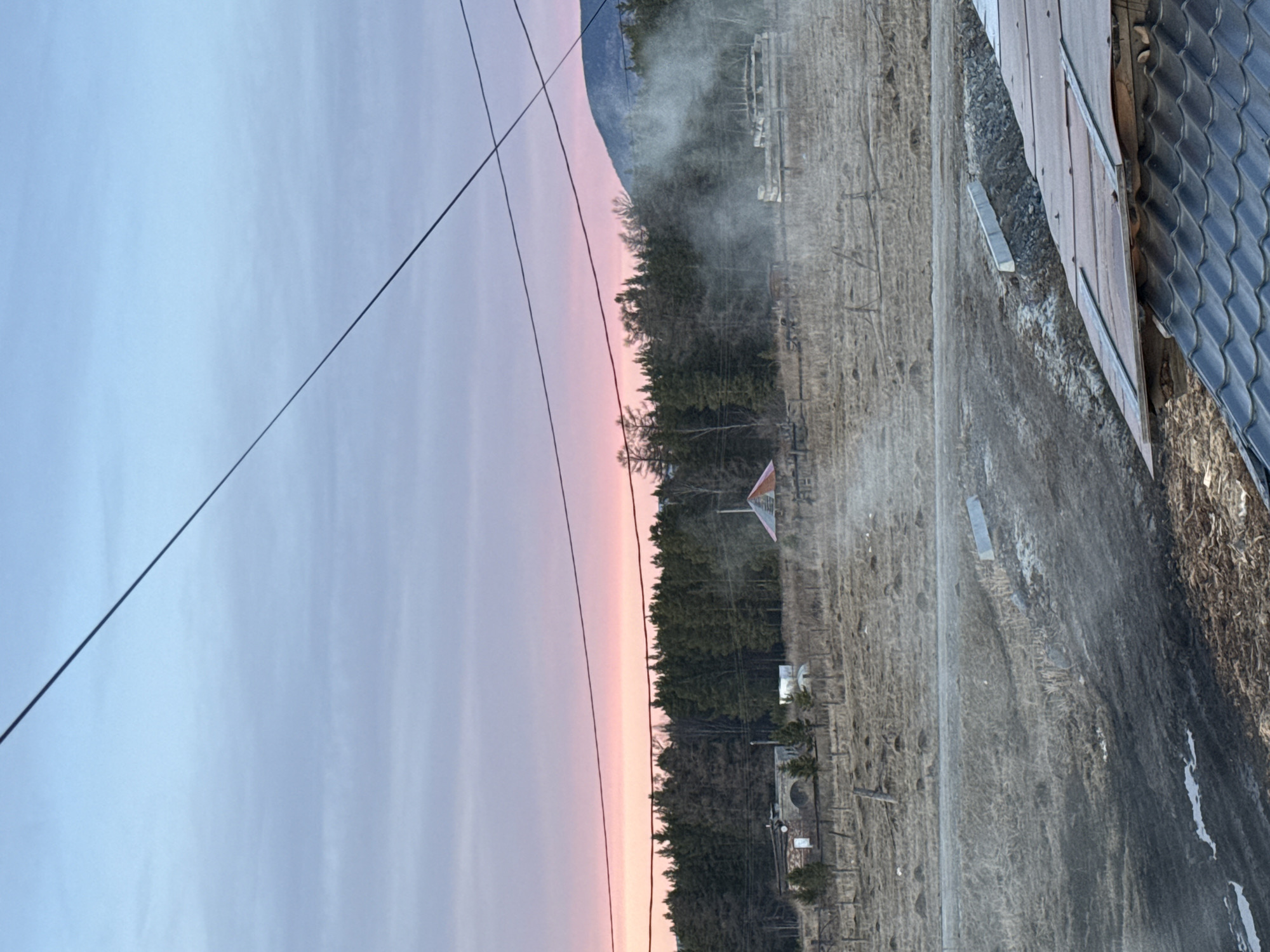 The image shows a landscape at dusk or dawn. In the background, a mountain range is visible, with a pink and purple sky overhead. A line of trees separates the sky from a field. In the foreground, there's a dirt road or path, and to the right, a wooden structure with a dark roof. Power lines cross the sky in the upper left. There's some haze or dust in the air, especially near the treeline. The image is rotated counter-clockwise. I cannot confirm the location is in Tyulyuk, Russia based solely on the visual information provided.