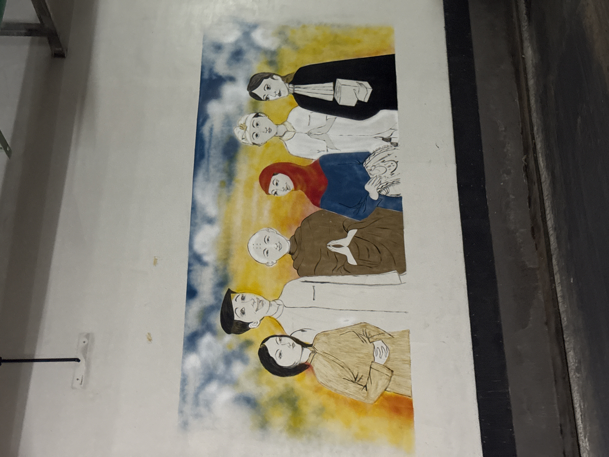 The image shows a mural painted on a white wall. The mural depicts six figures standing side-by-side against a backdrop of yellow and blue clouds. From left to right, the figures appear to represent different cultural or religious backgrounds. The first figure is a woman in a light brown robe. Next to her is a man in a white outfit. The third figure is a person in brown robes, possibly a monk, with hands pressed together in a prayer gesture. Following is a woman wearing a red hijab and a blue garment. Then there is a person in white attire with a head covering, also with hands pressed together. The last figure is a woman in a black robe and a white collar, holding a book. The mural is located in Kuta Selatan, Indonesia. The wall itself appears to be part of an interior space, possibly a hallway or corridor, as indicated by the hanging fixture at the top of the image. The lower part of the image shows a dark, textured surface, suggesting a floor or a lower wall section.