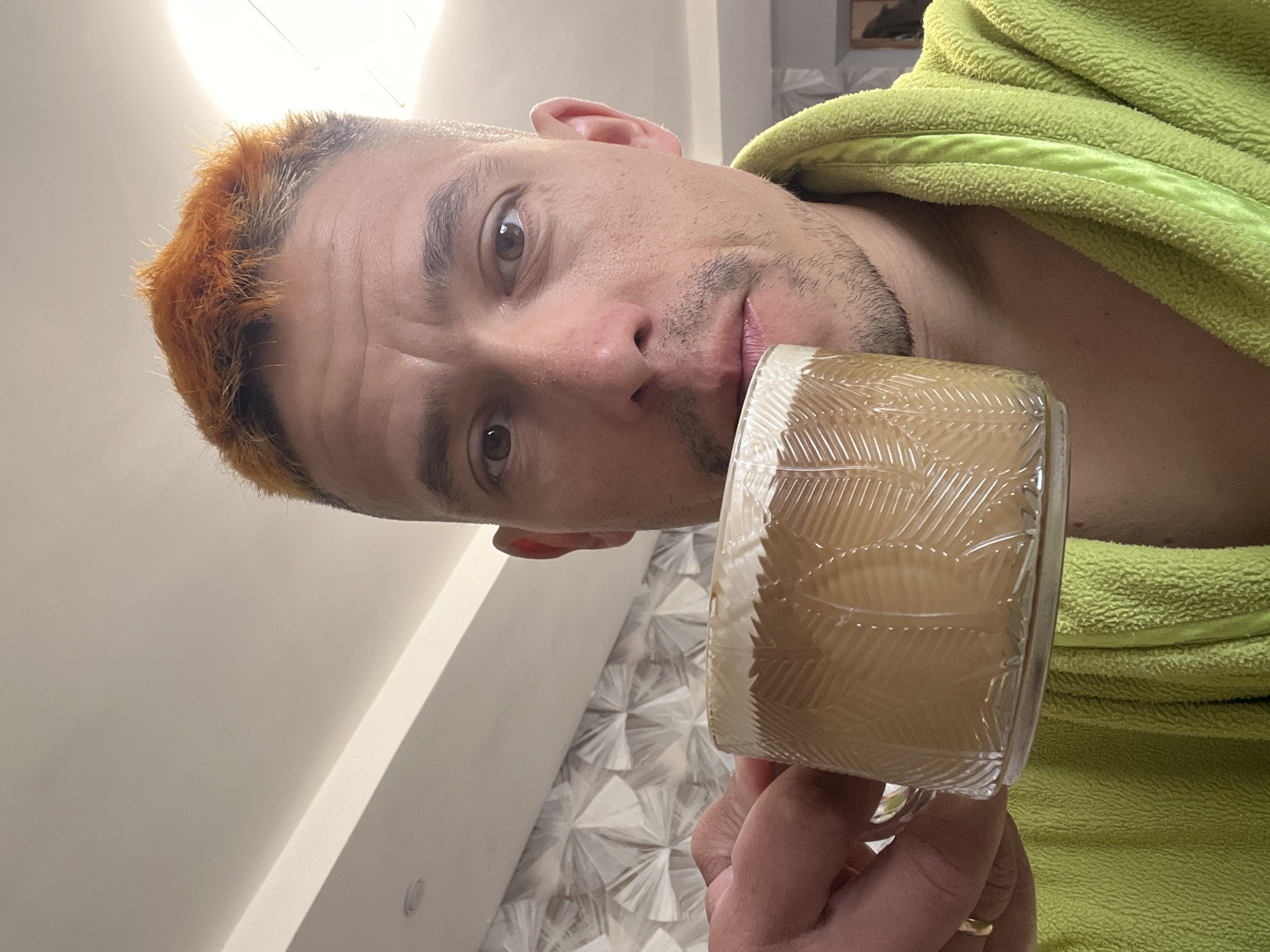 A man with dyed orange hair stands indoors holding a glass mug with a light brown beverage, potentially coffee or tea. He is wearing a light green terry cloth bathrobe. The interior features a textured wall pattern and standard ceiling lighting. There is no information to determine the location is specifically in the city of Chaikovsky, Russia.