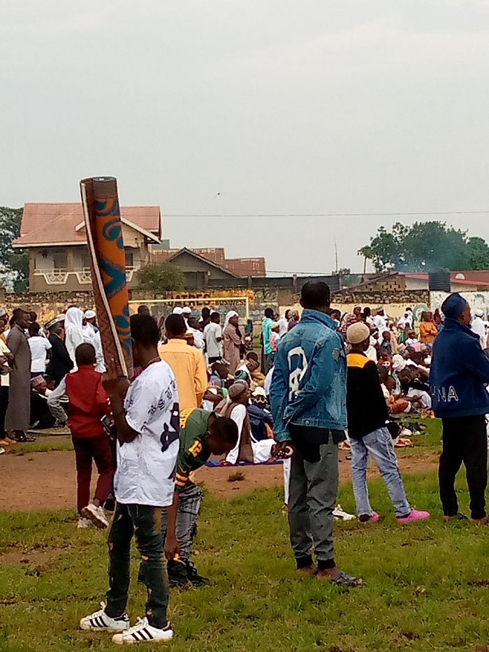 The image shows a gathering of people in an open field in Beni, Democratic Republic of Congo, located at coordinates 0.489 latitude and 29.464 longitude. The ground is mostly covered with grass, and there are a few buildings in the background. There are approximately 50 people in the scene, most of them are men and boys. They are all dressed in casual clothing. Many are sitting on the ground, while others are standing. A young man is standing in the foreground, holding a rolled-up rug or carpet. There are several people standing behind him, looking at the rug or the camera. The people in the background are blurred, and it is difficult to tell what they are doing. There appears to be a small amount of smoke in the background. The scene is peaceful and there is no evidence of any activity that could be considered dangerous or harmful.