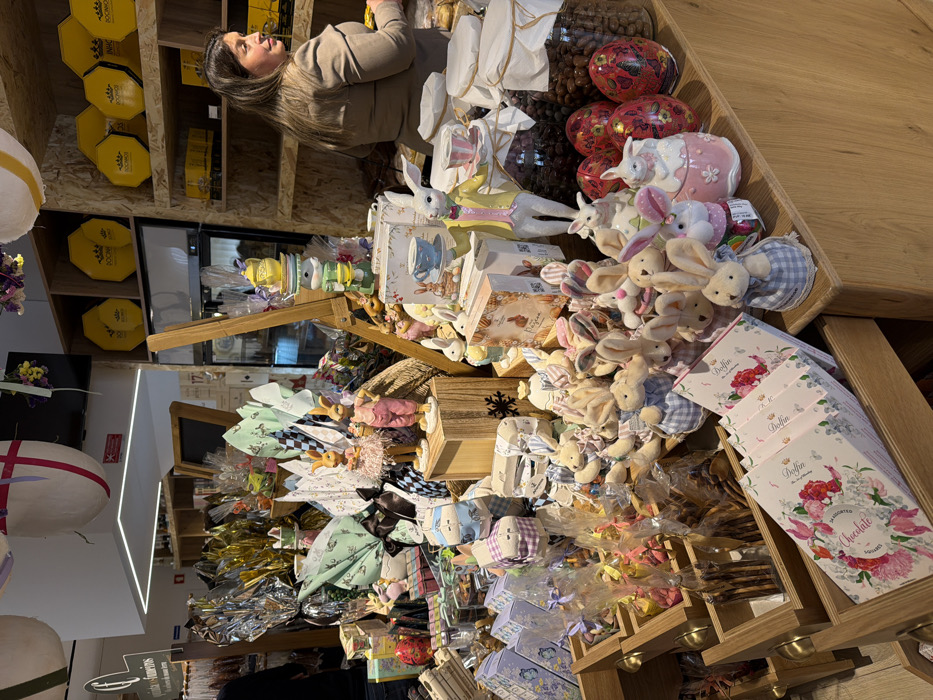This image depicts the interior of a confectionery or gourmet food shop located in Felgueiras, Portugal, densely arranged with Easter-themed merchandise.
**People:** One female individual is visible on the right side of the frame, standing behind a display counter. She has dark hair and wears a light brown, long-sleeved top. Her head is turned to her right, and her hands are partially visible, suggesting she is attending to items on the counter. A second person, largely obscured, is partially visible on the far left, facing away from the camera.
**Subjects/Objects:** The central focus is a large, multi-level display counter overflowing with Easter products. This includes numerous plush rabbit toys, many dressed in checkered blue and white fabric, and various ceramic rabbit figurines, some holding miniature teacups. Large, foil-wrapped chocolate eggs in gold, silver, and green are interspersed throughout the display, alongside smaller gift-wrapped items. Several decorative red and black patterned Easter eggs are visible on the right side of the display, adjacent to two large glass jars containing chocolate-covered nuts or similar confectionery. In the foreground on the left, wooden display drawers hold packaged sweets, including white boxes with pink floral designs labeled "Delfin Chocolate." A tall, stacked display of yellow ceramic teacups and a teapot, topped with a rabbit figurine, stands on a wooden easel-like structure. In the background on the right, wooden shelves display octagonal yellow boxes, some branded "BOCCA." A large, white egg-shaped decoration with red stripes hangs from the ceiling.
**Actions/Interactions:** The female individual appears to be working, possibly organizing or serving from the counter. No direct interaction between individuals is observed.
**Environment:** The interior space is brightly lit by ceiling fixtures, including a long rectangular LED light. The decor incorporates extensive use of wood for shelving and display units. The abundance of rabbit motifs, decorative eggs, and themed packaging indicates a festive, seasonal retail environment focused on Easter.