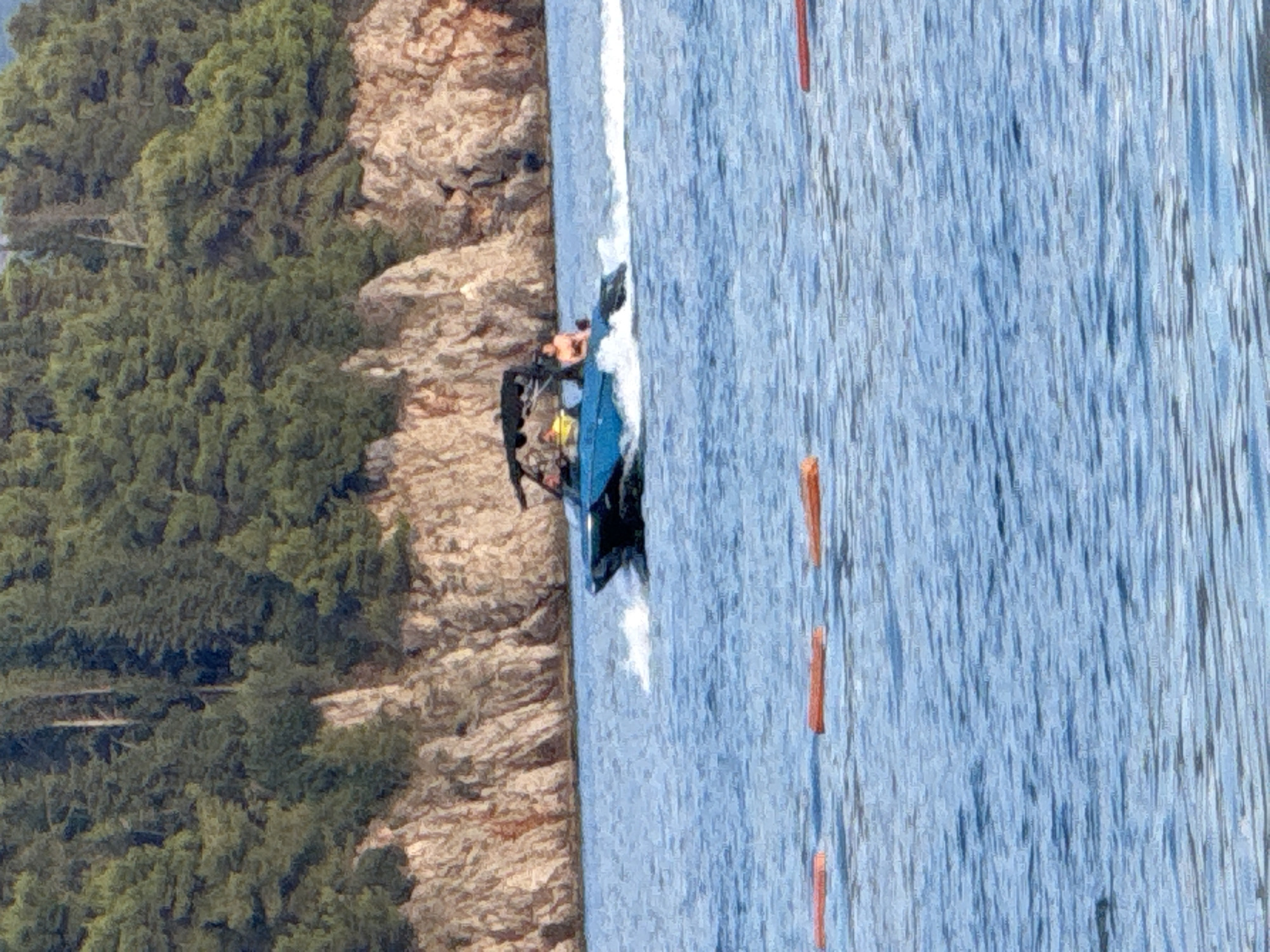 In this image, the scene is a body of water next to a rocky cliffside covered in dense green trees. The water is a bright blue with visible ripples and some white foam, suggesting movement. There are several orange buoys floating in the water, marking a path or area.
On the water, a blue and black personal watercraft, possibly a jet ski or a small boat, is visible. A person in dark clothing is seated on it. The watercraft appears to be kicking up a white spray, indicating it is moving or has recently moved at speed.
The cliffside is textured and layered, with a mix of light brown and grey rocks, interspersed with patches of green foliage. The trees are a deep, lush green and grow densely along the upper parts of the cliff.
The lighting suggests it is daytime, with the sun illuminating the scene and creating highlights on the water and rocks. No specific time of day or weather conditions beyond a bright day can be inferred. There is no visible text in the image. The overall impression is a recreational activity on a beautiful natural waterfront.