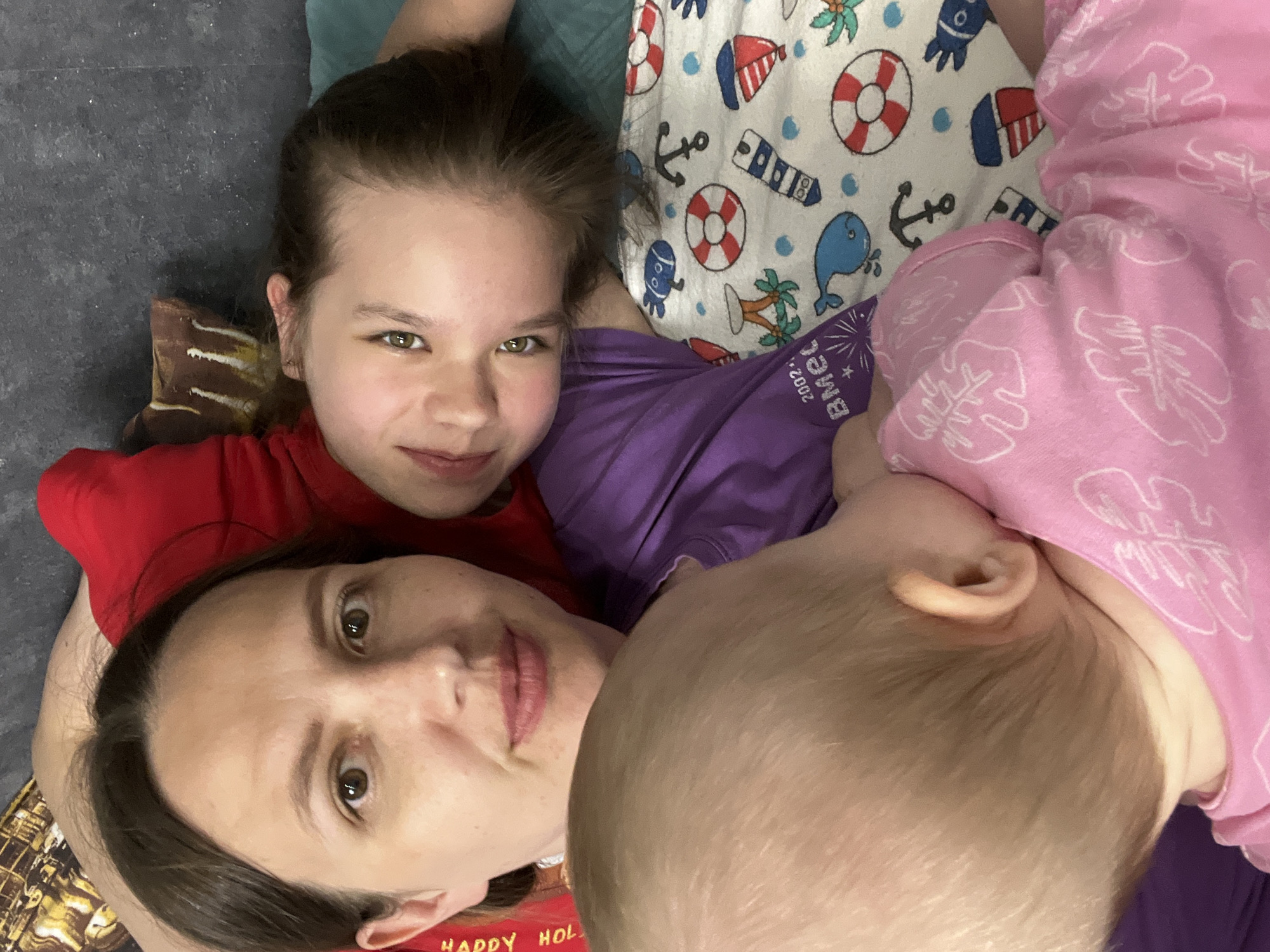 A close-up, eye-level photograph shows three individuals and various objects. A woman with brown hair and eyes, wearing a red shirt with "Happy Holidays" partially visible, is positioned at the bottom left. A girl with brown hair and green eyes, wearing a purple shirt, is above the woman. To the right of the girl is a baby wearing a pink patterned pajama. A blanket with nautical-themed images of anchors, life preservers, whales, and palm trees is behind the girl. The surface in the background appears to be a dark gray color. Based on the text "BMec 2002" visible on the purple shirt, and a search of the internet, this image may be related to a person associated with the city of Chaikovsky, Russia. The individuals appear to be lying down close together, possibly taking a selfie.