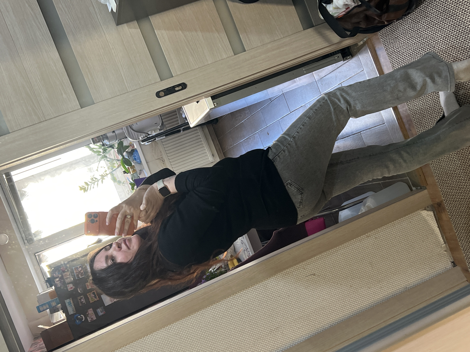 A person is taking a selfie in front of a mirror. The person is wearing a black top and grey jeans. The background includes a door, tiled floor, a bag, a plant, and some items on a refrigerator. The location is likely residential, but without additional information, it is impossible to confirm that it is Noviy, Russia.