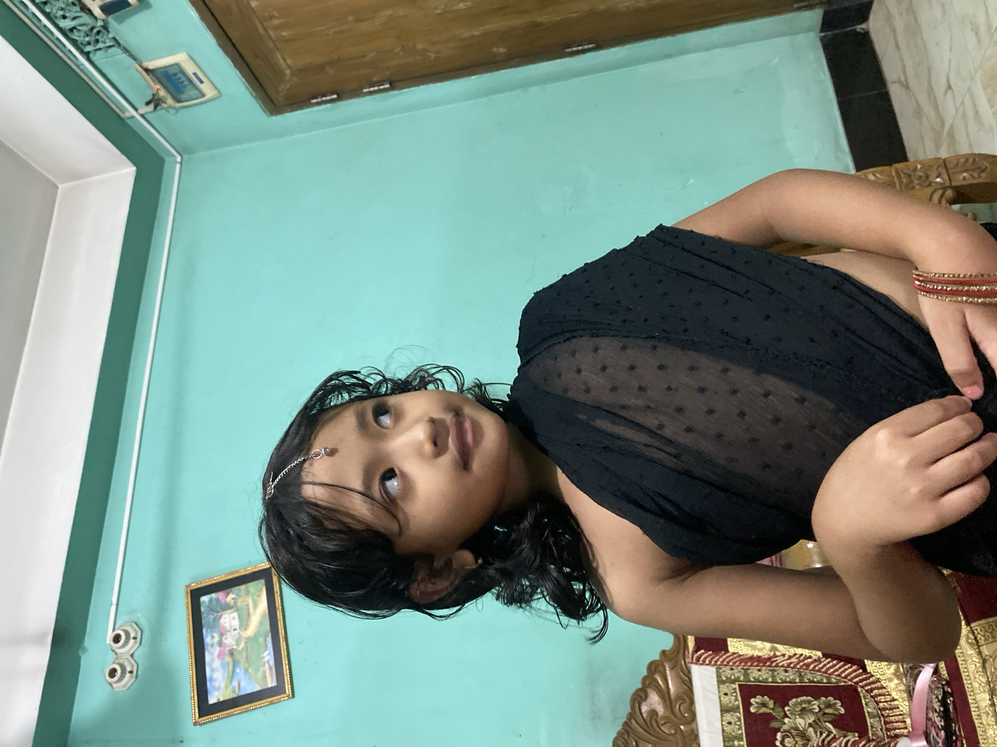 The image shows a young girl dressed in a black sari, sitting on a wooden chair. She is wearing a headpiece with a small dangling charm. The girl has dark, curly hair and is looking to her right with a slight smile. She is wearing gold bangles on her right wrist. Behind her, a teal-colored wall is visible, with a framed picture of a landscape hanging on it. To the right of the girl, a wooden door is present. The scene appears to be indoors, possibly in a residential setting within Dhaka, Bangladesh.