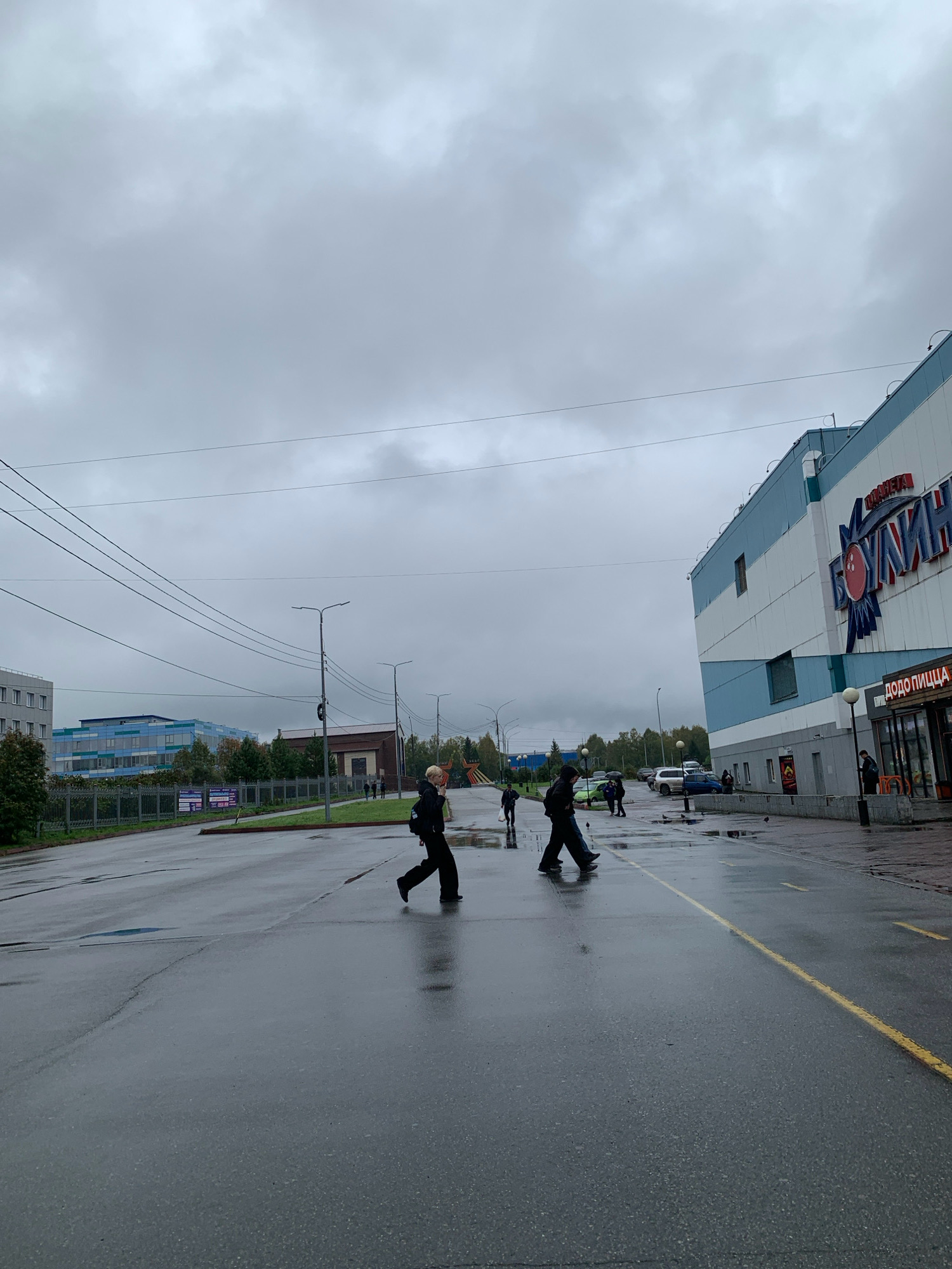 The image depicts a street scene in Prokopyevsk, Russia, on an overcast day. The foreground is dominated by a wet asphalt road with yellow lane markings, indicating it is a pedestrian crossing. Two individuals are crossing the street from right to left in the mid-ground. One person is wearing dark clothing and a backpack, while the other is also dressed in dark attire. To the right of the street is a large, modern building with blue and white signage that includes Cyrillic text and a bowling pin graphic. A smaller storefront with "DODO PIZZA" in Cyrillic text is visible adjacent to the larger building. In the background, across the street, are several buildings, including a blue and glass structure, and a brick building. Trees line the background, and utility poles with numerous wires are present. The overall atmosphere is grey and slightly damp, suggesting recent or ongoing rain.
