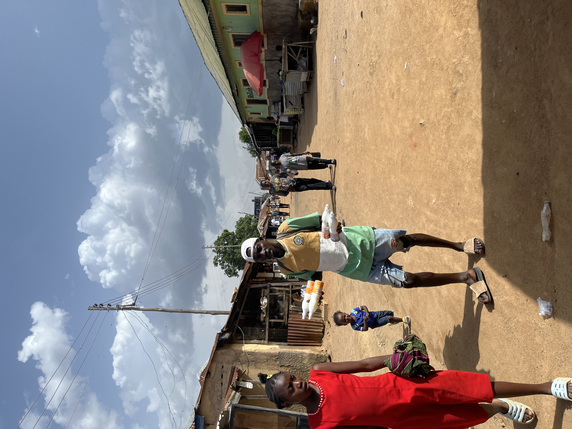The image depicts a bustling street scene in Jos, Nigeria, under a bright, partly cloudy sky. The time of day appears to be late morning or early afternoon, given the strong shadows cast by the buildings and people. The setting is a dusty, unpaved street lined with small shops and market stalls, suggesting a vibrant local marketplace.
The primary subjects are several people. In the foreground, a young girl in a red dress and white sandals is looking towards the left. Behind her, a man wearing a green and yellow t-shirt, ripped denim shorts, and sandals stands holding two bottles of liquid. He appears to be a vendor or seller. Further back, other individuals can be seen walking or standing near the shops, and a young child in a blue shirt is lying down on the ground.
The activity appears to be the daily commerce of the marketplace. The man with the bottles is likely showcasing his products. The overall atmosphere seems lively and active. Notable details include the overhead power lines, typical of many Nigerian towns, and the corrugated metal roofing on some of the stalls. The buildings are simple structures with weathered paint.
There is some visible text on a building in the background that reads "Shiloh". The lighting and shadows suggest fair weather, possibly with some dramatic cloud formations contributing to the sky's appearance. The image conveys a sense of everyday life in a Nigerian community.