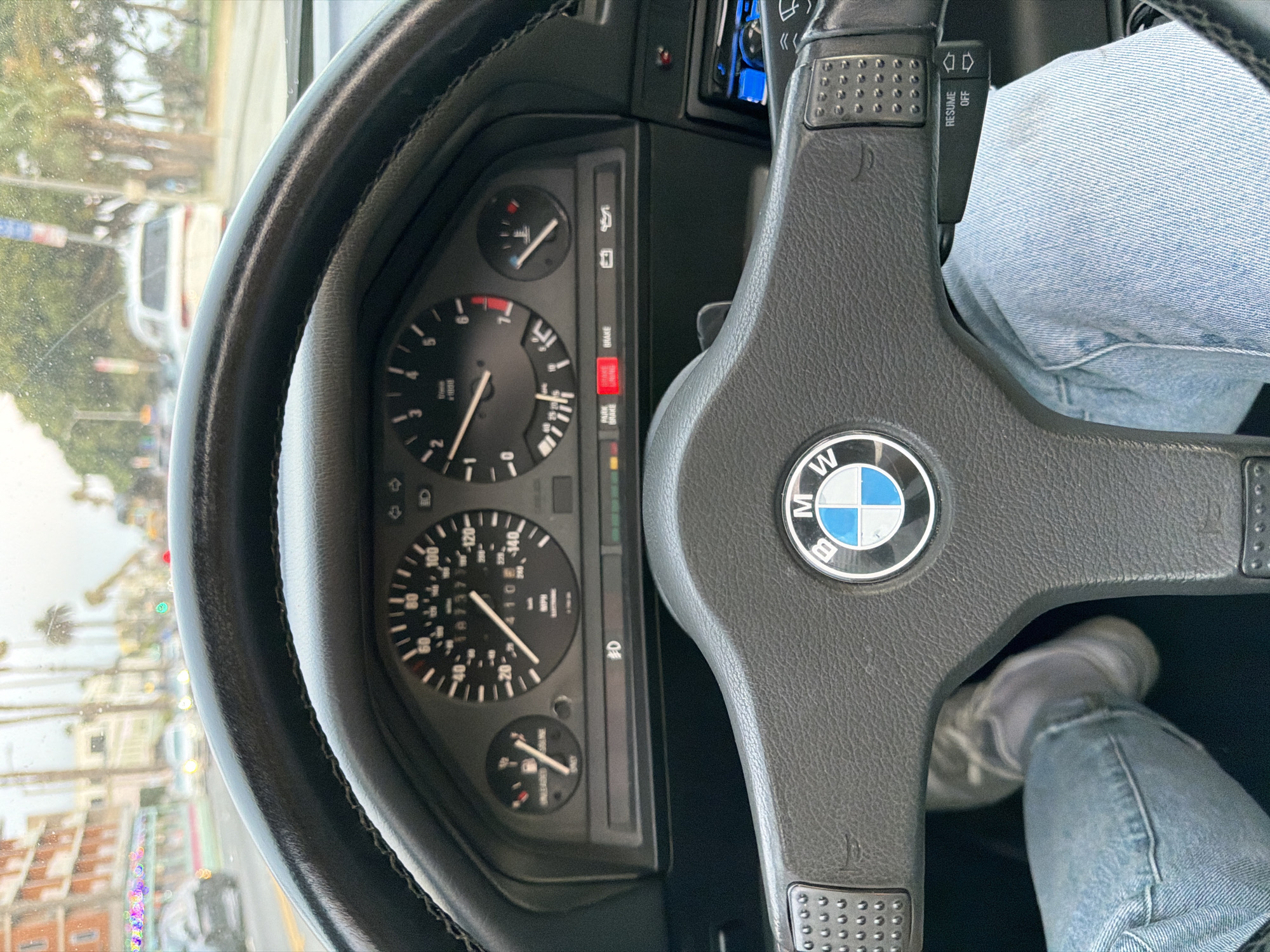 The image is a first-person perspective from inside a BMW, looking at the dashboard and the road ahead through the windshield. The driver's legs, clad in light blue jeans and grey athletic shoes, are visible in the lower part of the frame. The steering wheel, featuring the BMW logo, is prominent. The dashboard displays a speedometer, tachometer, fuel gauge, and temperature gauge. Several warning lights are illuminated, including "PARK BRAKE" and "BRAKE". The exterior view shows a street scene with buildings, palm trees, and other vehicles, suggesting an urban environment. The lighting and atmospheric conditions, with some moisture on the windshield, indicate it may be overcast or raining. The architecture of the buildings and the presence of palm trees are characteristic of a Southern California city like Santa Monica.