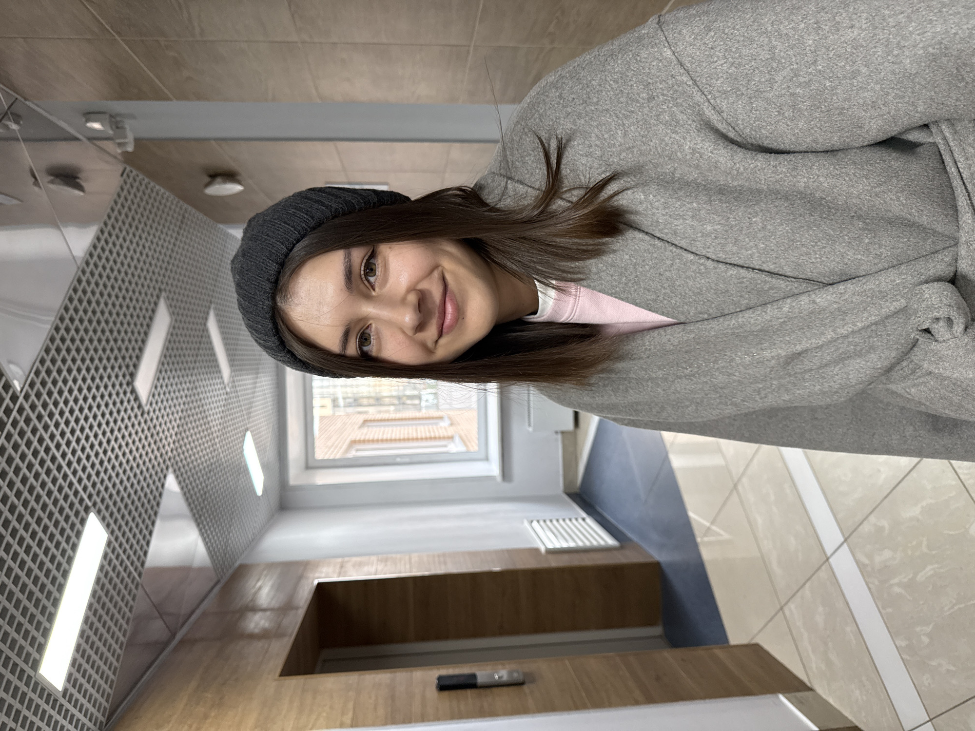 A young woman is posing in what appears to be an indoor hallway. The woman has brown hair, brown eyes, and is wearing a dark gray beanie, a pink shirt, and a gray wool coat with a belt. She is smiling slightly at the camera. The hallway has a light-colored tiled floor with dark gray insets. The wall on the left has a distinctive geometric grid pattern, and illuminated rectangular lights are embedded in the wall. A wooden panel is on the lower part of the wall to the right. In the background, a window reveals the facade of a brick building. The presence of brick buildings and modern interior design suggests that the location could be a renovated building in an urban area like Krasnoyarsk, Russia. A pen is on the wooden panel.