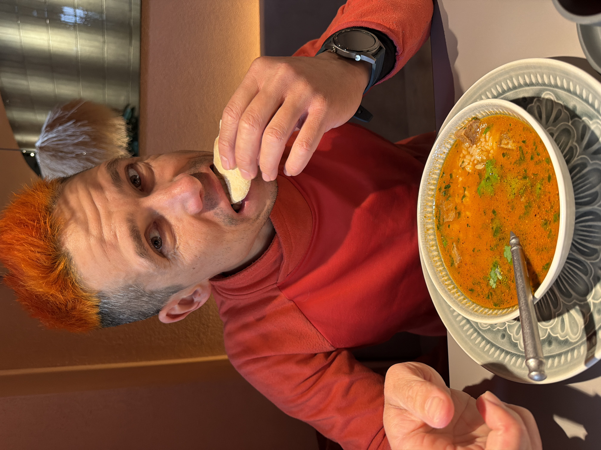A man with orange hair and a red long-sleeved shirt is seated at a table. He is holding a piece of bread to his mouth with his left hand, while his right hand rests on the table. On the table is a bowl of orange soup with green herbs and rice, along with a spoon. The soup bowl rests on a gray plate with a circular pattern. The man is wearing a smart watch on his left wrist. The setting appears to be indoors, with a tan-colored wall in the background and a blurry reflection of an outdoor scene in the upper left corner. There are no indicators of this picture having been taken in Chaikovsky, Russia.
