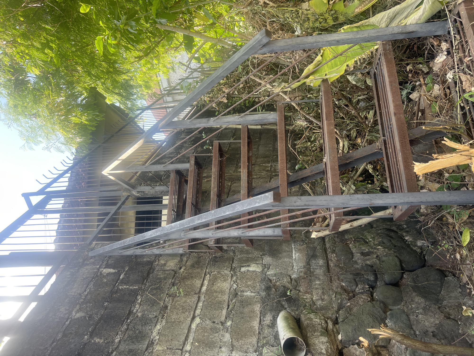 An exterior, low-angle, medium shot captures a rusty, metal staircase with a railing ascending alongside a weathered, grey concrete block wall on the left. The staircase is positioned in an outdoor environment with abundant greenery and foliage to the right, suggesting a tropical setting, likely in Denpasar, Indonesia. Sunlight filters through a dense canopy of lush green trees above, casting a bright, slightly hazy glow on the scene. The staircase itself shows signs of age and exposure, with visible rust and grime on its metal treads and supports. A white PVC pipe with some green moss is attached horizontally to the lower portion of the concrete wall on the left. At the bottom of the frame, debris such as fallen leaves, twigs, and a piece of broken wood are scattered on the ground. The wall is constructed from irregularly shaped, dark grey stones at its base, transitioning to larger concrete blocks with visible mortar lines higher up. The background behind the staircase reveals a building with a tiled roof and a window. The overall impression is one of an aged, somewhat neglected outdoor area in a warm, verdant climate.