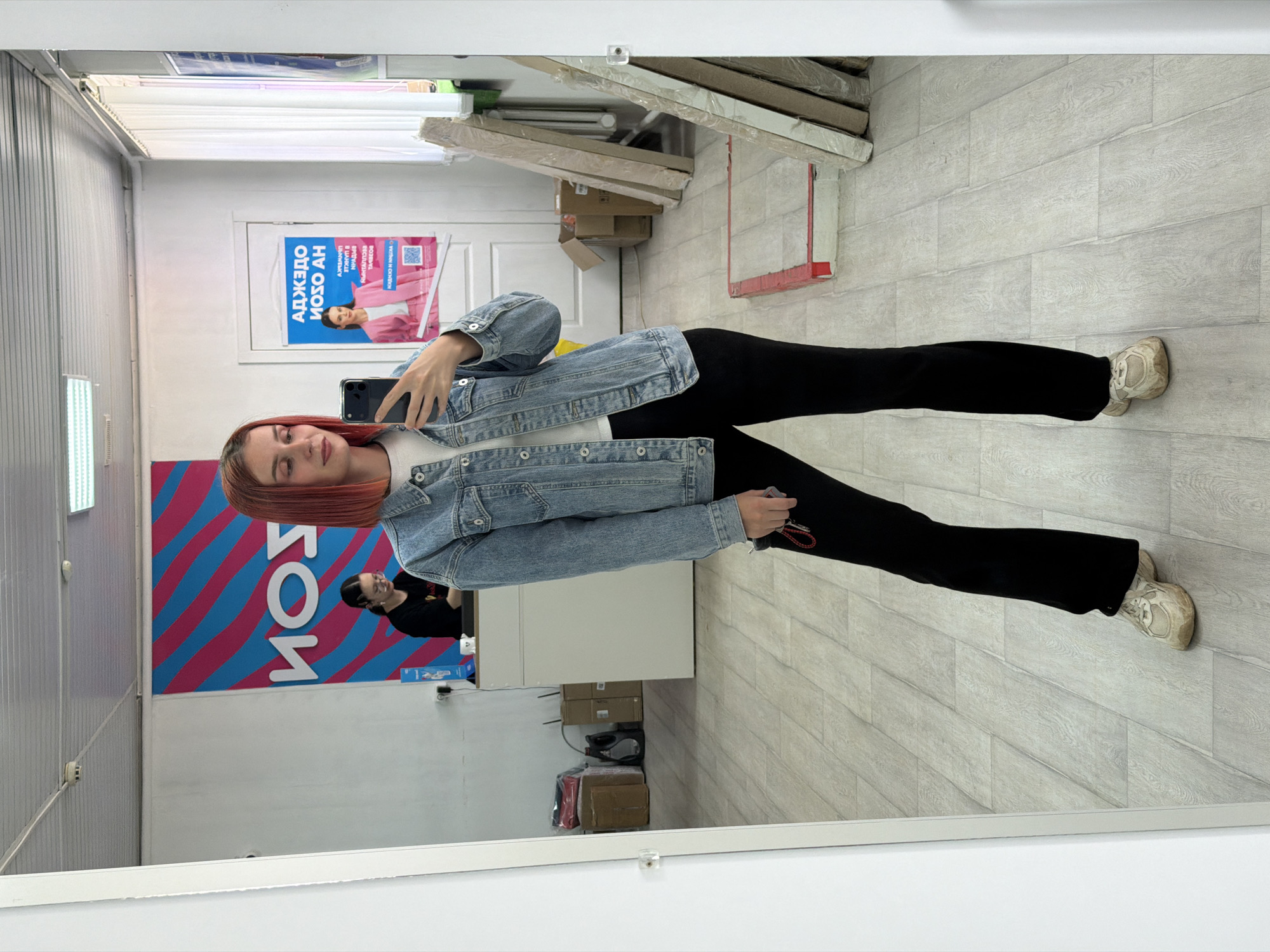 The image shows a woman taking a selfie in a mirror. She is wearing a denim jacket, black pants, and white sneakers. She has reddish-orange hair. The reflection in the mirror reveals a store interior. Visible in the background are shelves, a promotional poster with Russian text ("АДЖЕДО ИОЗО АН"), and other store items. Another individual wearing black clothes is partially visible in the mirror's reflection. The mirror's edge is visible at the bottom of the frame. The floor appears to be tiled with a light-colored wood-grain pattern. Based on the text on the wall and the general appearance, the location is in Chaikovsky, Russia.