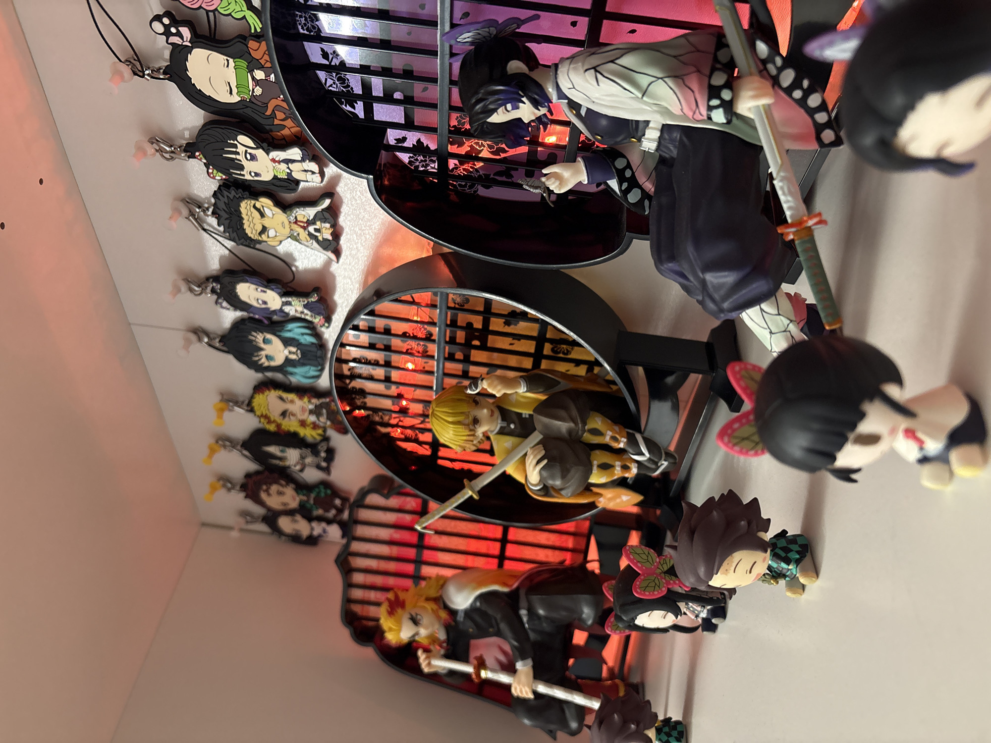 The image shows a display of figurines and keychains featuring characters from the anime series "Demon Slayer". The figurines are arranged in a tiered display case. The keychains are hung along the side of the display case. The lighting is a combination of natural light and colored lights, possibly from within the display case. The background is a plain white wall. The location is not Plovdiv, Bulgaria, since there is nothing in the image to indicate a connection to that city.