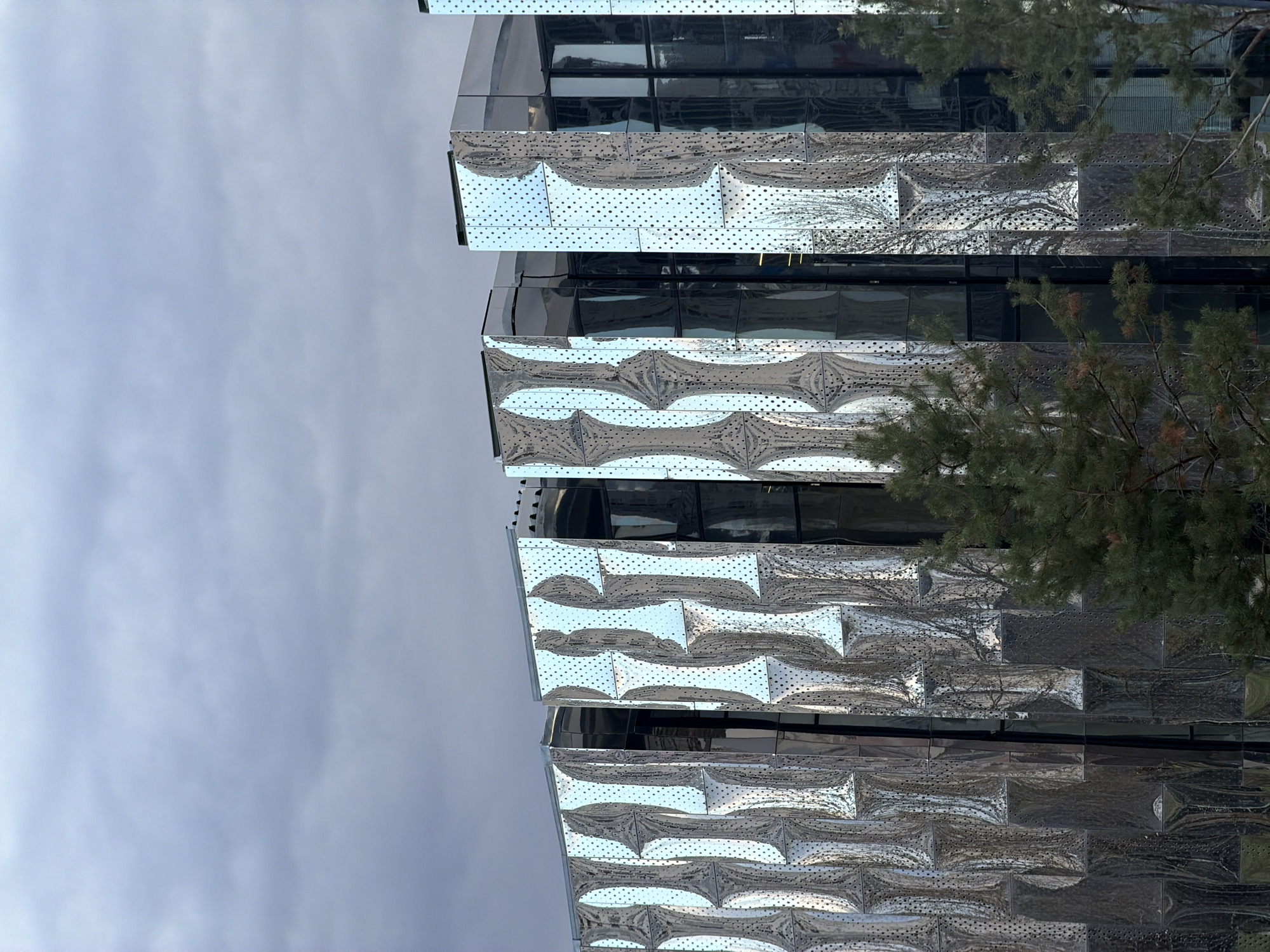 A low-angle shot of a modern building, with its exterior adorned with reflective, patterned metal panels, is seen against a cloudy sky. The building appears to be situated in Khimki, Russia. The metal panels have a textured, almost brick-like arrangement and create shimmering reflections of the surroundings. Green foliage from nearby trees is visible on the side, adding contrast to the building's metallic facade. The building's architecture suggests a contemporary design with sharp edges and reflective surfaces.