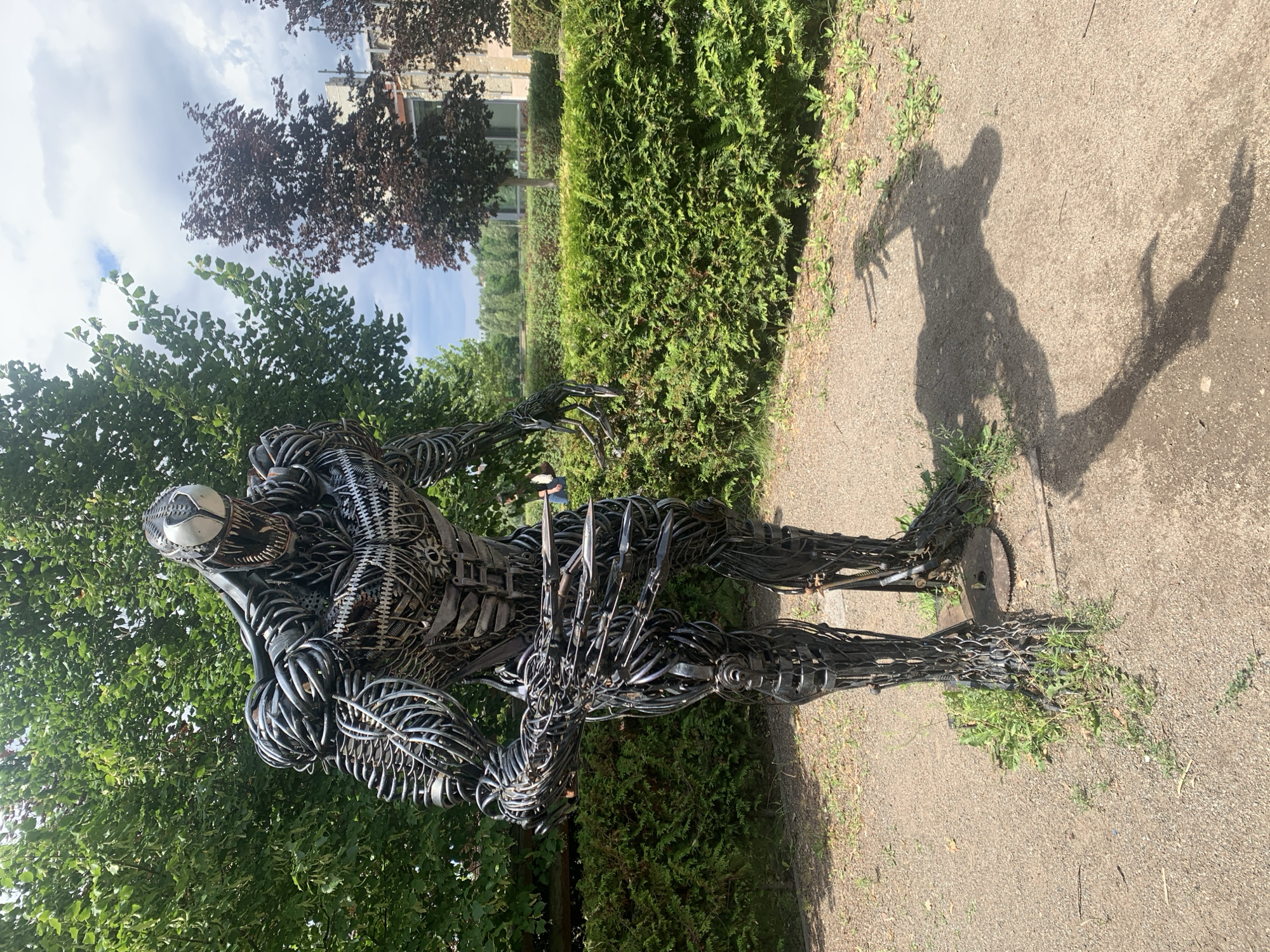 A full-body, frontal shot shows a large, dark metal sculpture of the comic book character Venom standing outdoors. The sculpture is intricately crafted from various metal components, resembling a biomechanical creature with sharp claws and a menacing grin. It is positioned on a gravel path, with patches of green grass growing around its base. Behind the sculpture, a well-maintained green hedge forms a backdrop, and beyond that, trees with lush green and dark red foliage are visible. In the distance, a modern building with large windows can be seen. The sky above is partly cloudy with patches of blue. The sculpture is casting a prominent shadow on the ground to its right, which also displays its clawed hands and menacing posture. This sculpture is located in Bucha, Ukraine.