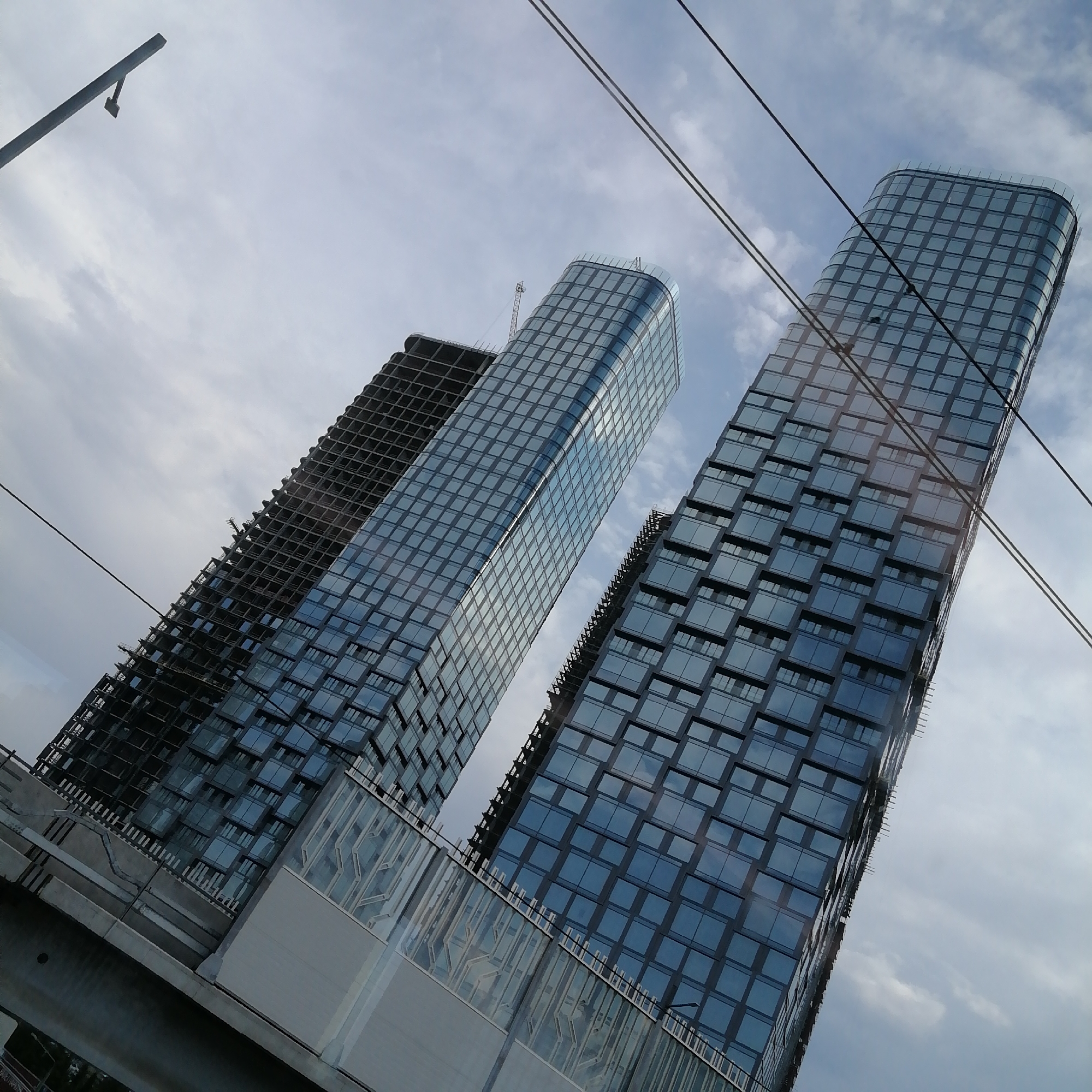 A low-angle shot captures a cluster of modern skyscrapers against a cloudy sky. The buildings are under construction, with one tower appearing unfinished, its concrete skeleton exposed. The other two towers are clad in glass panels that reflect the sky and clouds. Overhead, power lines crisscross the frame, and a streetlamp is visible in the upper left corner. The scene is tilted, giving a dynamic and slightly disorienting perspective.