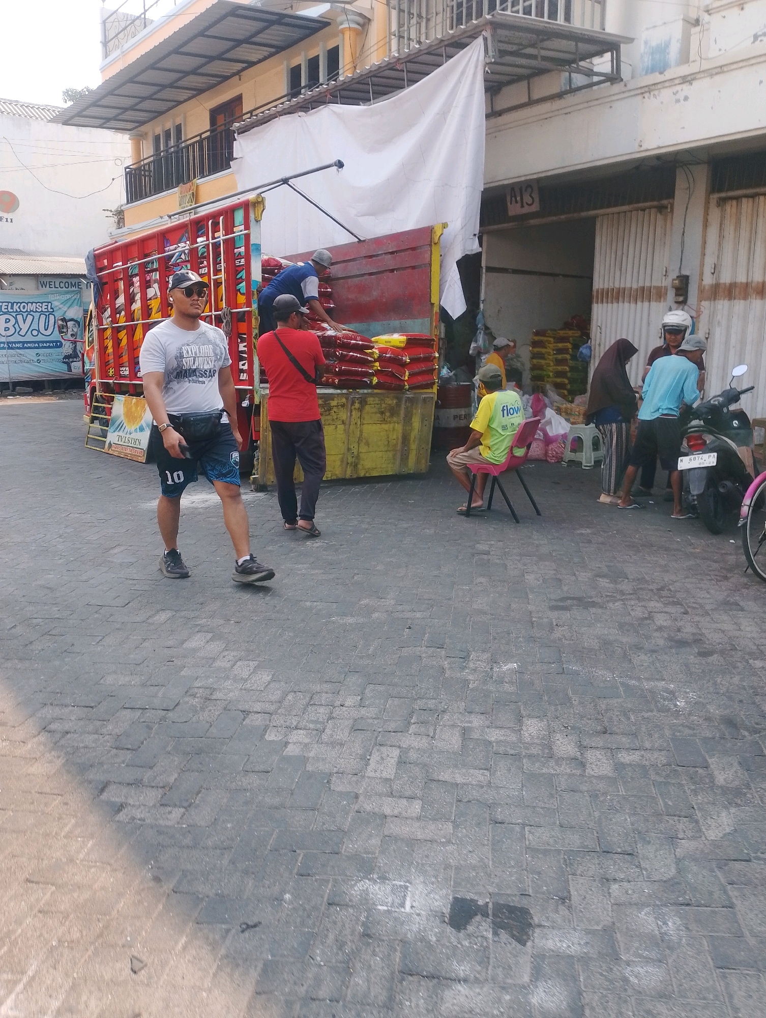 The image depicts a street scene in Probolinggo, Indonesia, with a focus on activity around a large red truck laden with sacks of goods. In the foreground, a man wearing a grey t-shirt, black baseball cap, and black shorts with a blue design is walking on a paved cobblestone street. He has a black fanny pack around his waist and is wearing athletic shoes.
Behind him, the red truck is the central element. Two men are actively engaged with the cargo. One man, wearing a red shirt and dark pants, is standing by the truck, and another man in a blue shirt and grey cap is partially visible behind him, appearing to be assisting with the sacks.
Further back, on the right side of the frame, a group of people are gathered near a building with a corrugated metal facade. One person, wearing a bright yellow t-shirt with "flow" printed on it, is seated on a red plastic chair. Another individual in a blue shirt is standing next to a motorcycle. A woman in a dark headscarf is also present in this group. Sacks of goods are stacked in the background, both on the truck and within the building's entrance. The environment appears to be a commercial or market area, given the presence of the truck and goods. The buildings have a visible level of wear, typical of urban environments in Indonesia.