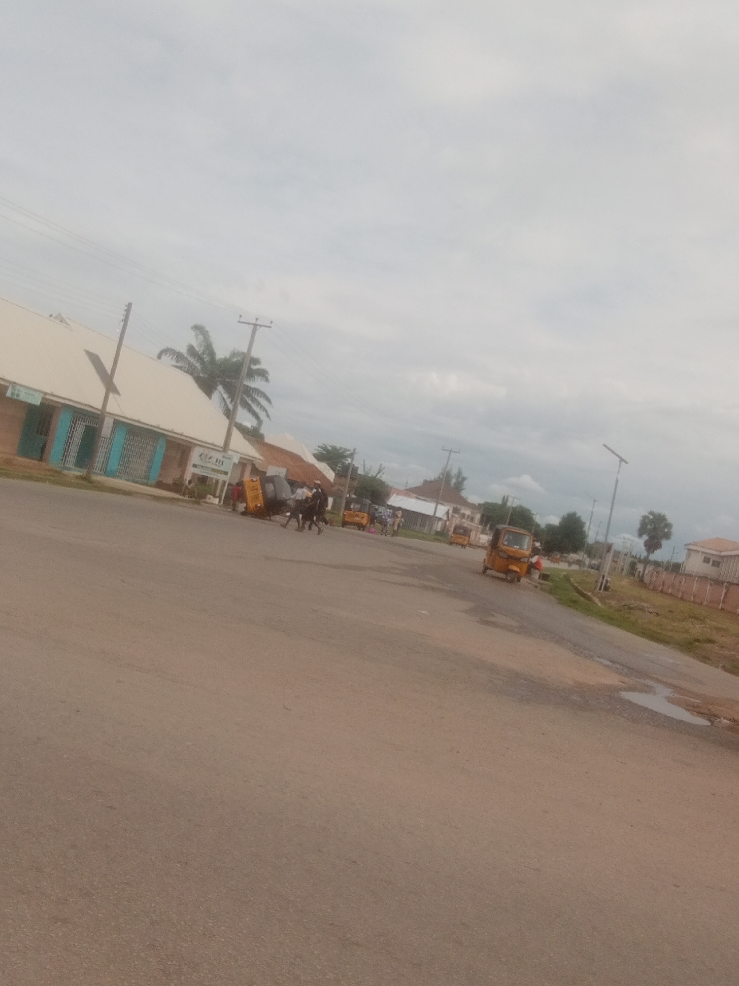 The image depicts a street scene in Jalingo, Nigeria, under an overcast sky. The foreground is dominated by a paved road, with patches of dampness and a small puddle suggesting recent rain.
Along the left side of the road, a row of low-rise buildings lines the street. These buildings have light-colored, sloped roofs and appear to house businesses, indicated by barred windows and an entrance with light blue trim. A sign with green text and a logo is visible on one of the buildings. Further down the street, more buildings are visible, some with tiled roofs.
Several three-wheeled vehicles, commonly known as "keke-napep" in Nigeria, are present on the road. One yellow keke-napep is parked near the right side of the frame, while another is in motion further down the street. A group of people are gathered around a keke-napep in the middle ground, with some individuals walking or standing.
A palm tree is visible behind the buildings on the left, and scattered trees and vegetation are present throughout the scene, particularly on the right side of the road. Utility poles with power lines stretch across the sky, a common sight in urban and semi-urban areas. The overall impression is that of a typical day in a Nigerian town, with ongoing daily activities. The cloudy weather suggests it might be morning or late afternoon.