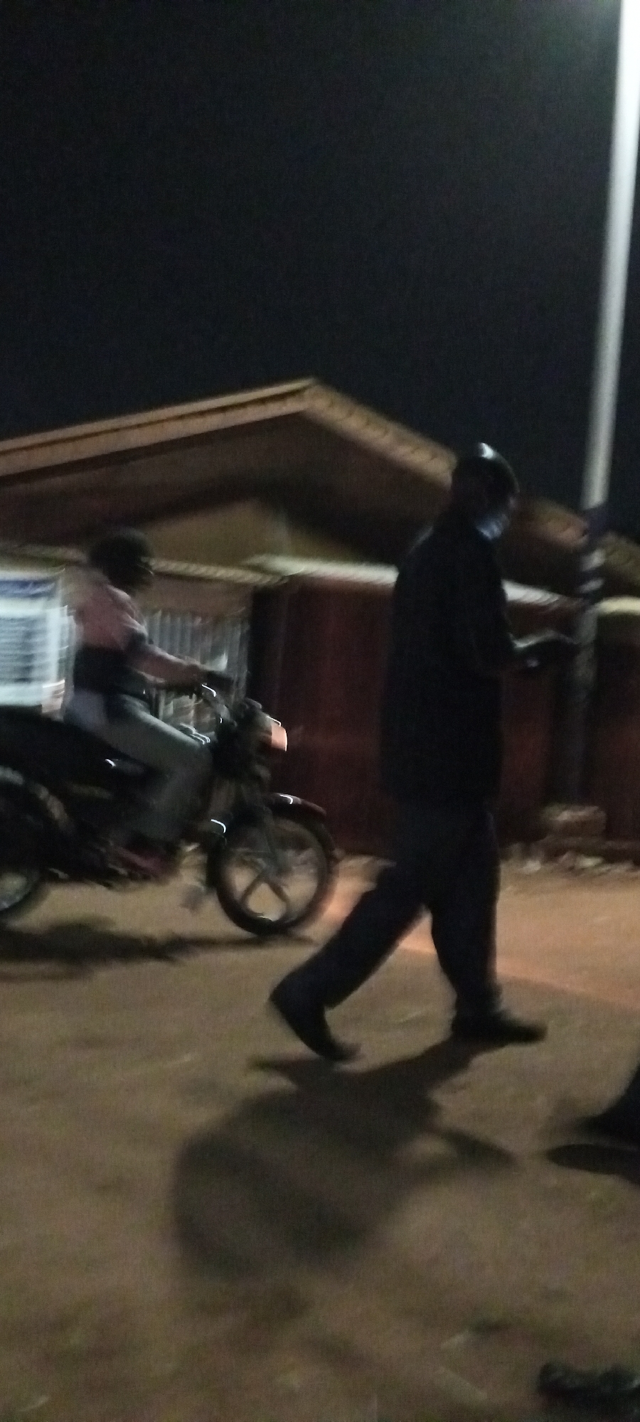 The image shows a street scene at night in Sabon Tasha, Nigeria. A person is riding a motorcycle on the left side of the image, while another person is walking on the right side. There is a building in the background with an overhang. A light pole is visible on the right side of the image. The street appears to be unpaved.